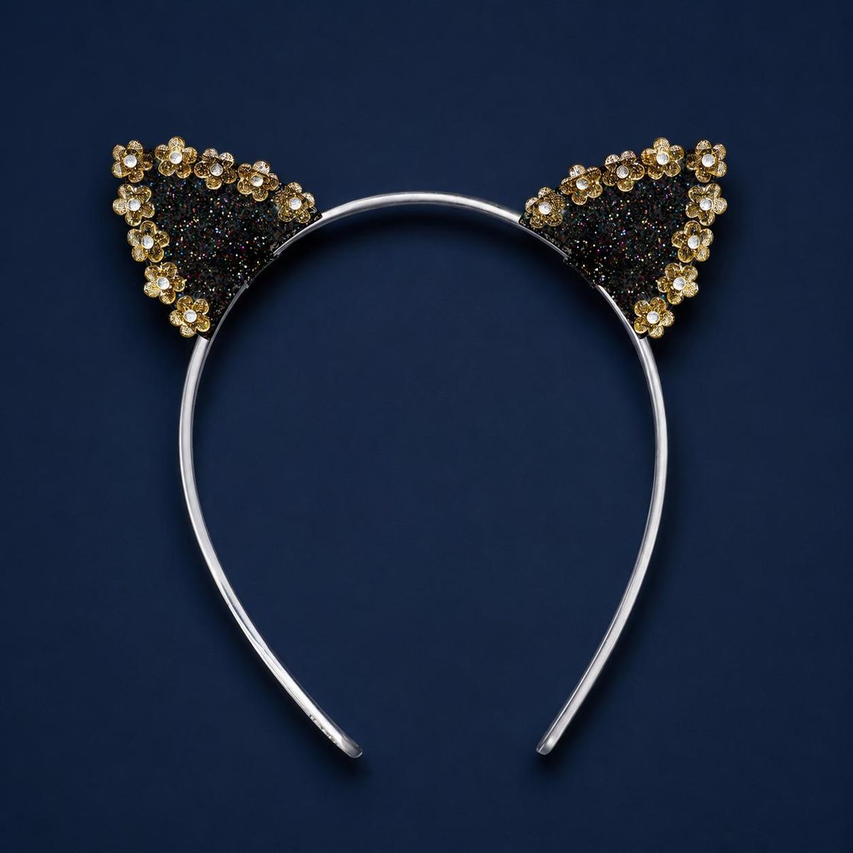 A cat-ear headband decorated with black glitter and small gold and white flower-shaped rhinestones, shown against a dark navy blue background.
