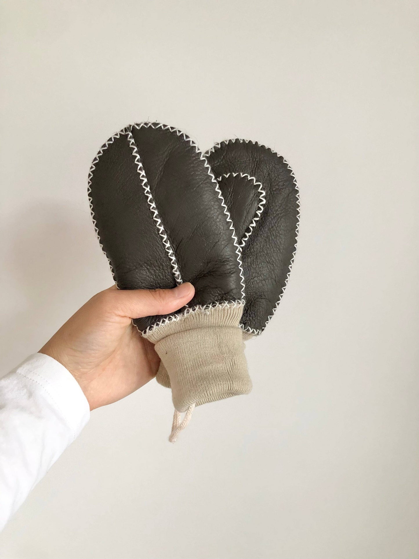Hand holding a pair of dark gray leather mittens against a white background