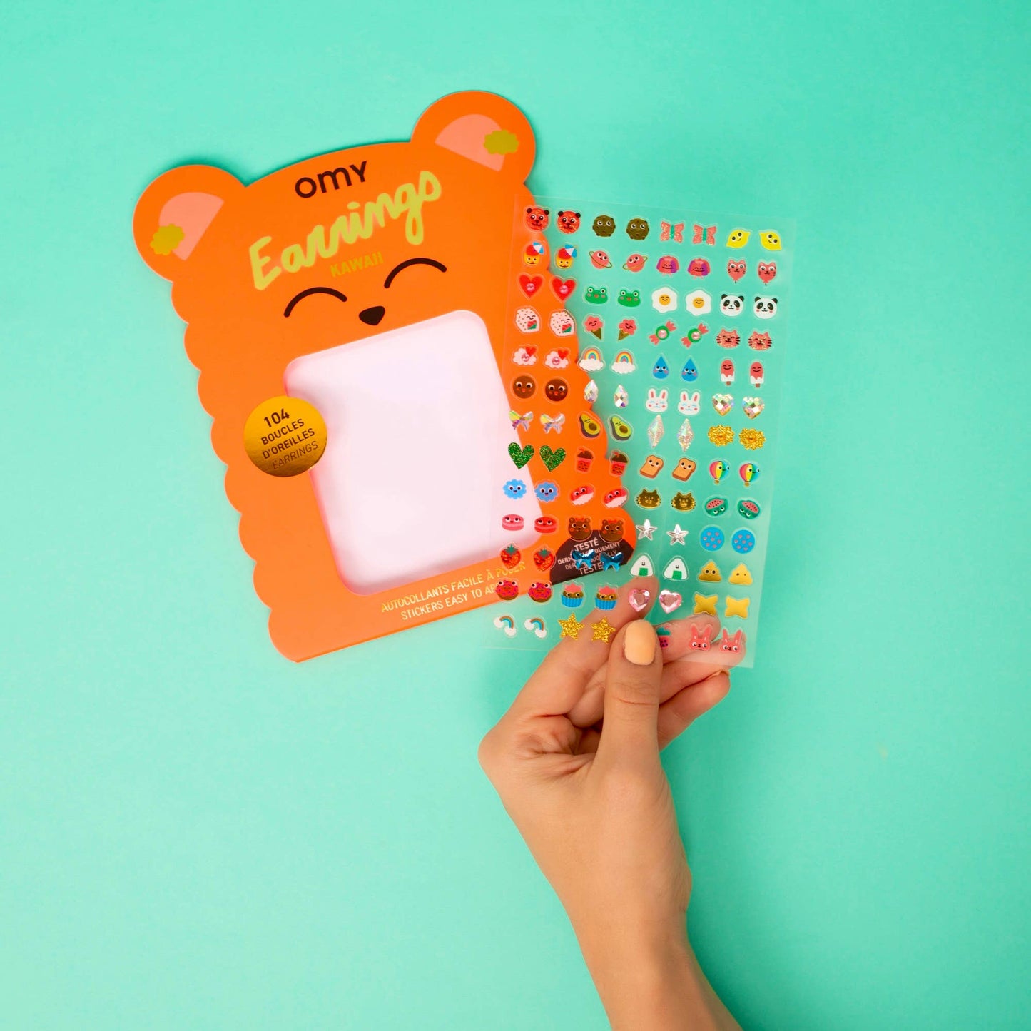 Hand holding a clear sheet of colorful earrings with an orange packaging on a teal background
