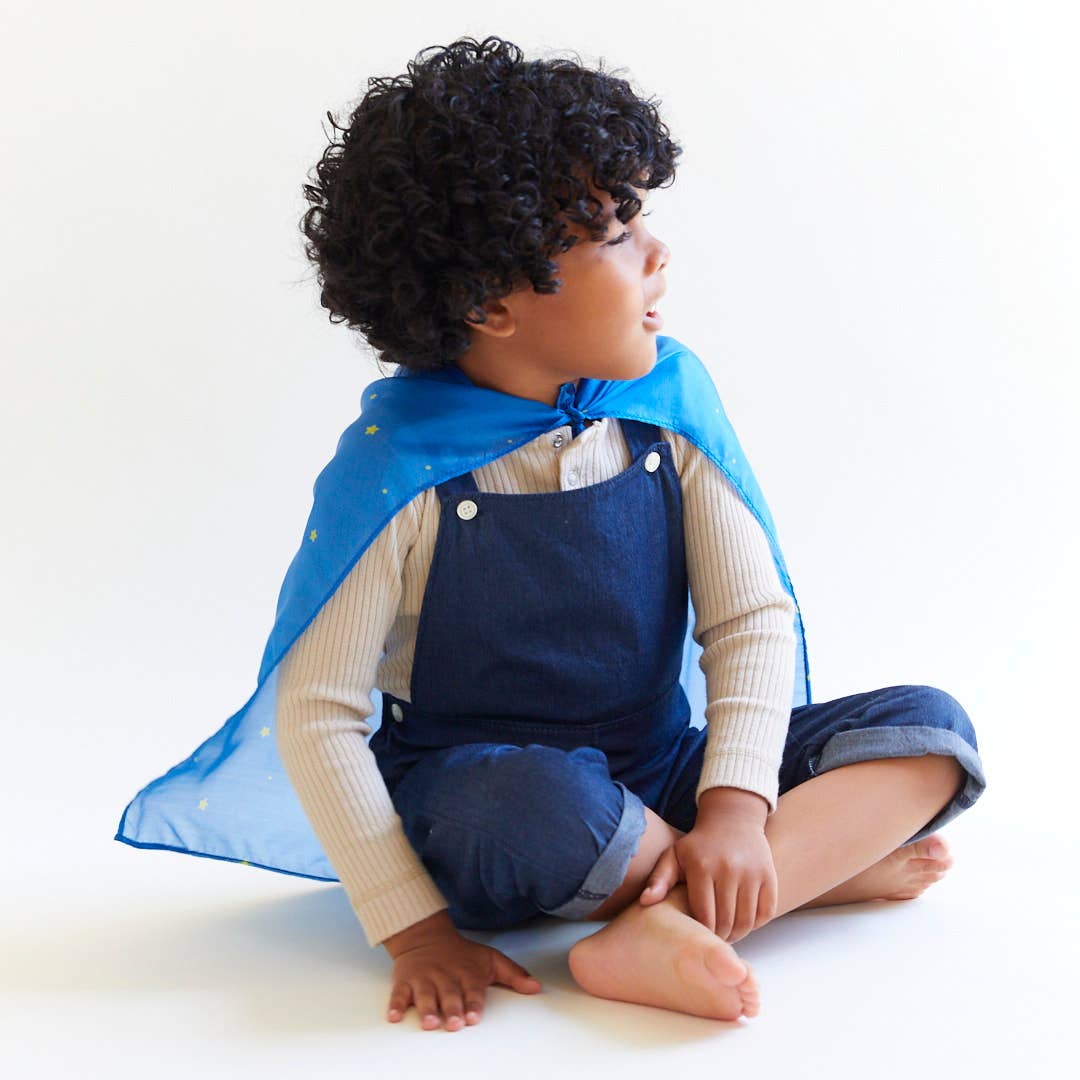 Child wearing a blue cape and denim overalls sitting on a white background