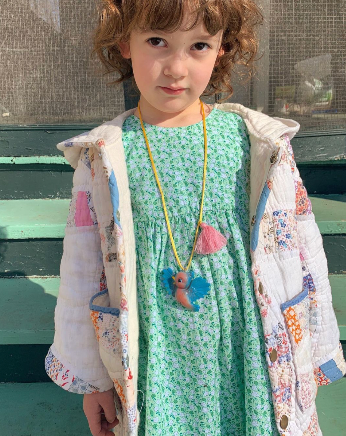 A young child wearing a mint green floral dress under a white patchwork quilted jacket, accessorized with a yellow cord necklace featuring a blue and pink fairy charm.