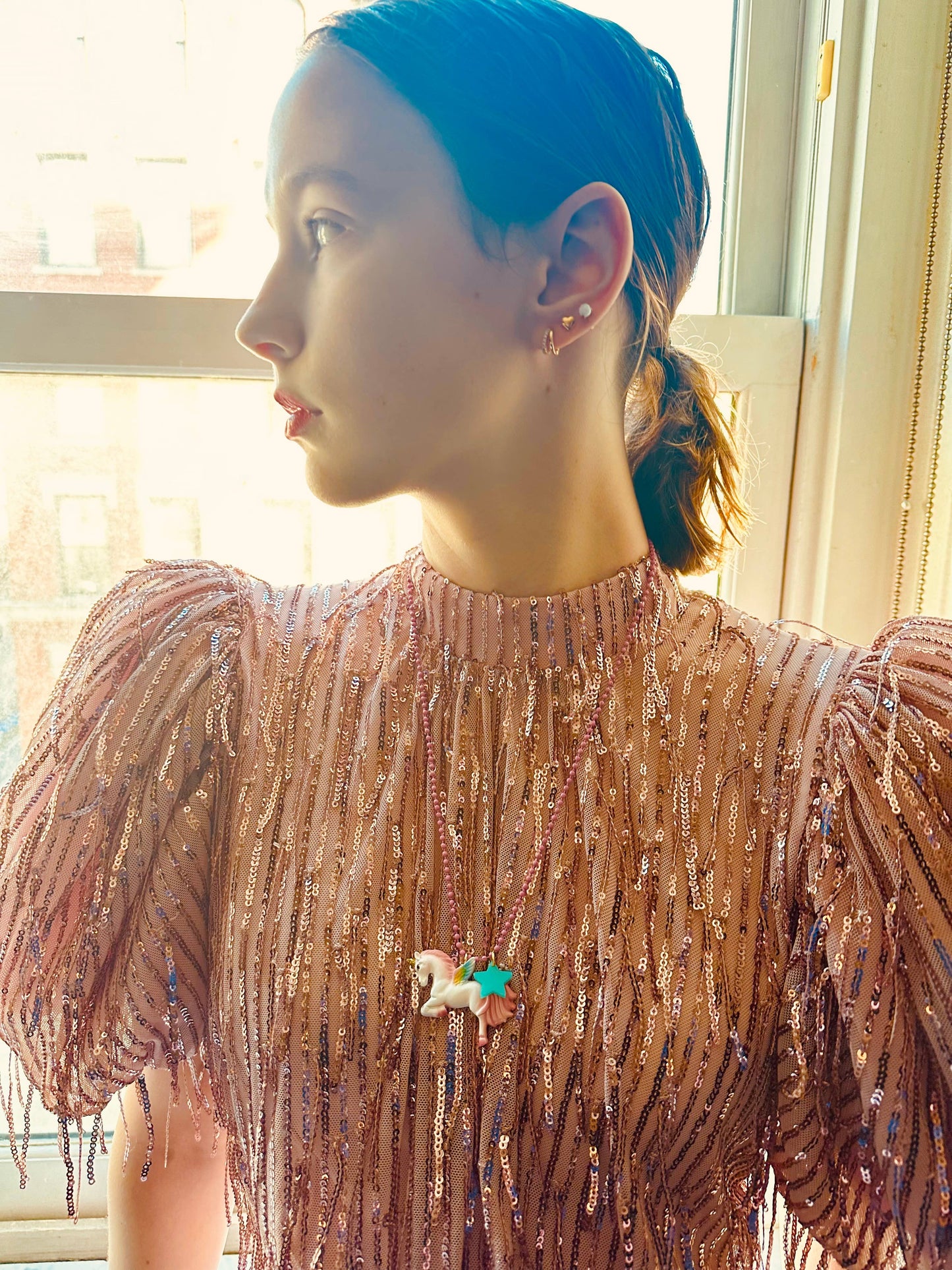 Person wearing a sparkling pink top with a unicorn necklace, standing in front of a window.