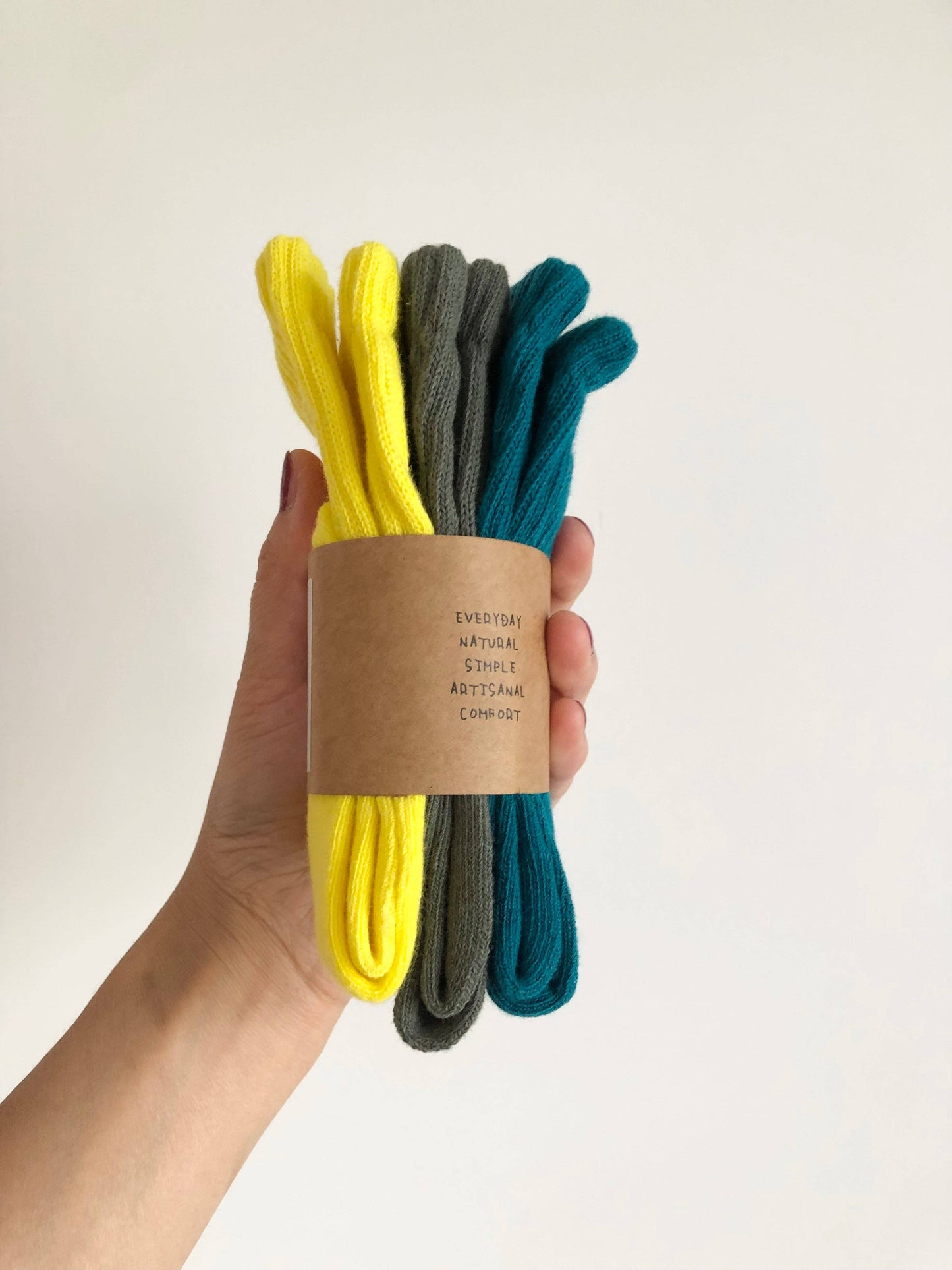 Hand holding a bundle of three pairs of socks in yellow, gray, and blue with a cardboard sleeve on a light background.