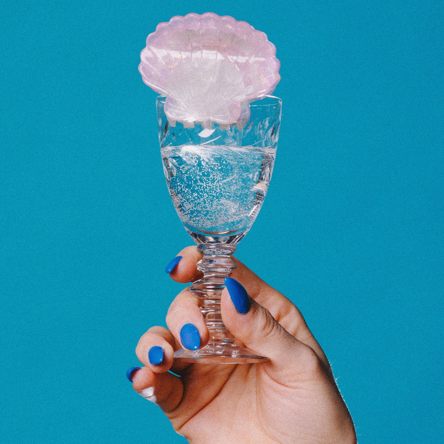Hand holding a small glass with a pink shell-like object against a blue background