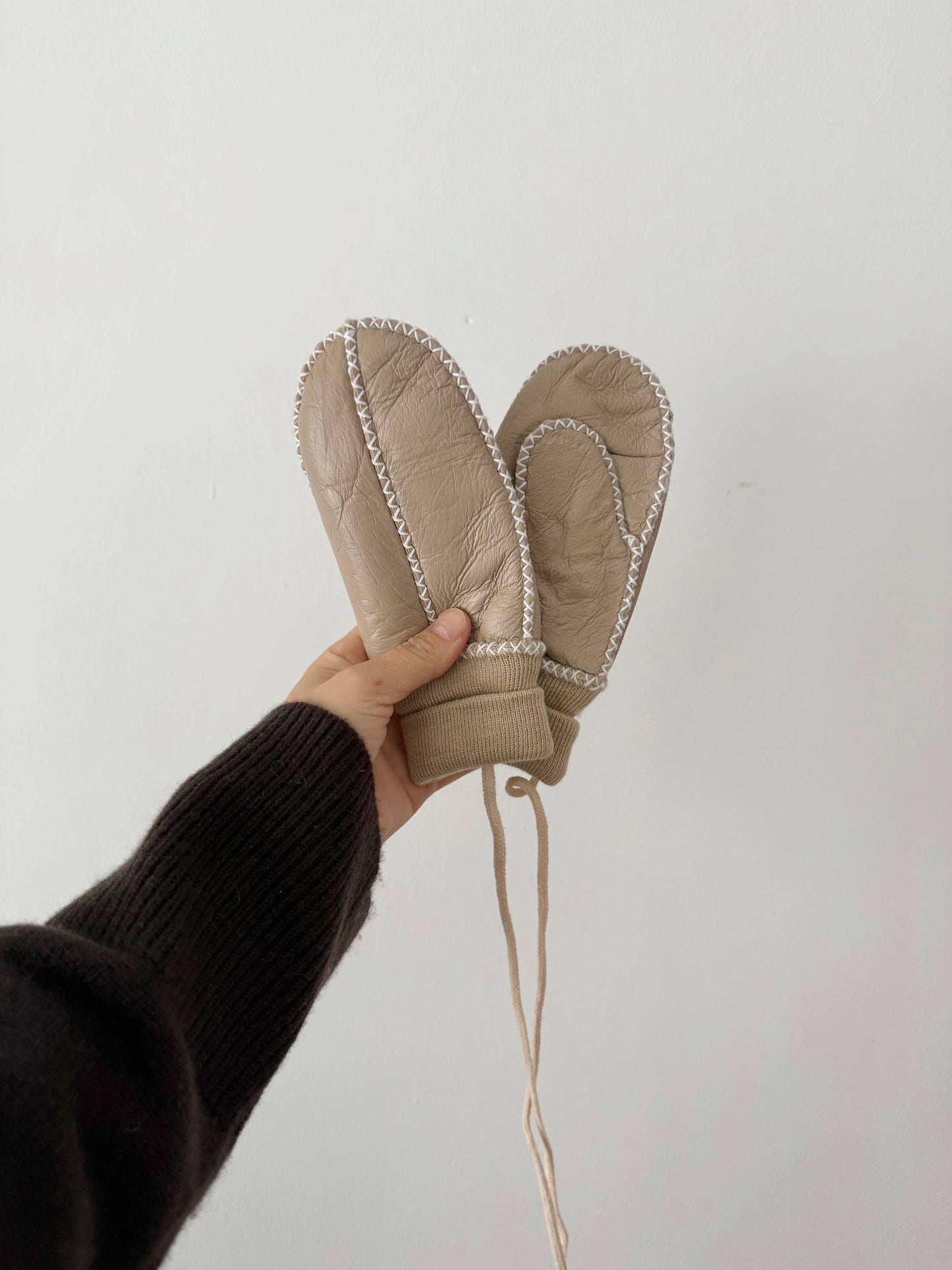 Hand holding a pair of beige leather mittens against a white background