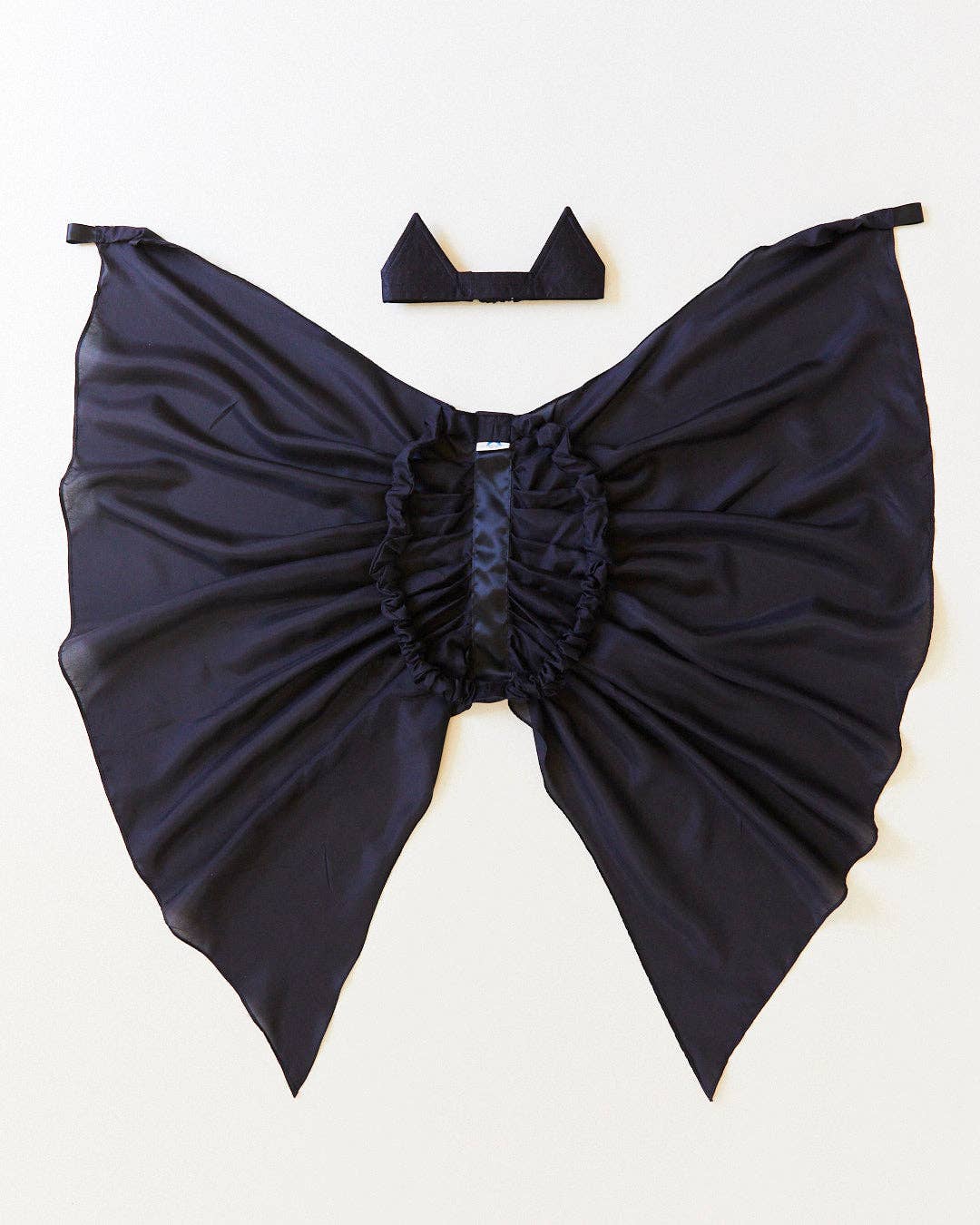A black bat-wing costume accessory with a gathered center and a small matching black headpiece, laid flat on a light off-white background.