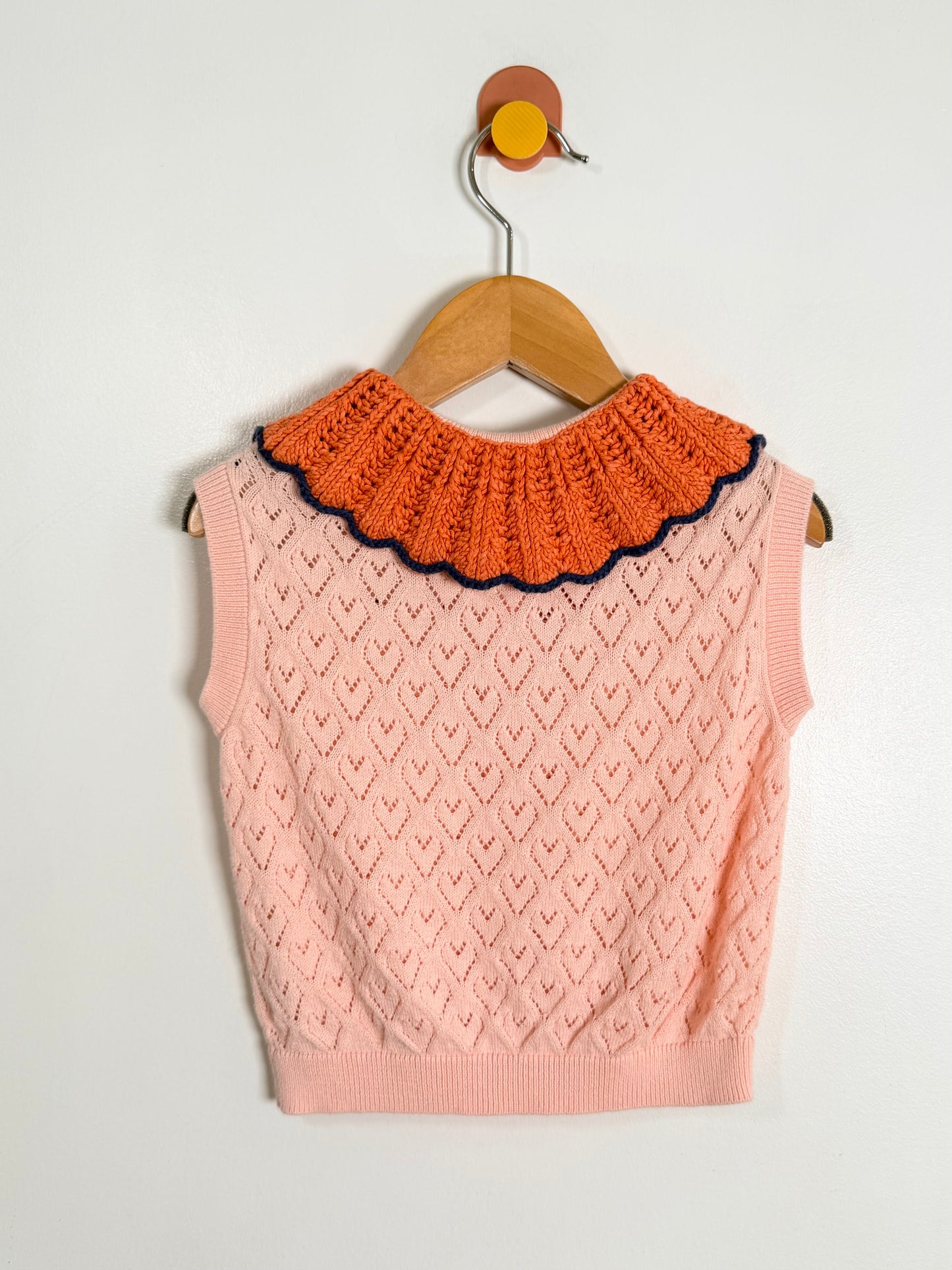 Pink knitted vest with orange collar on a wooden hanger against a light gray background