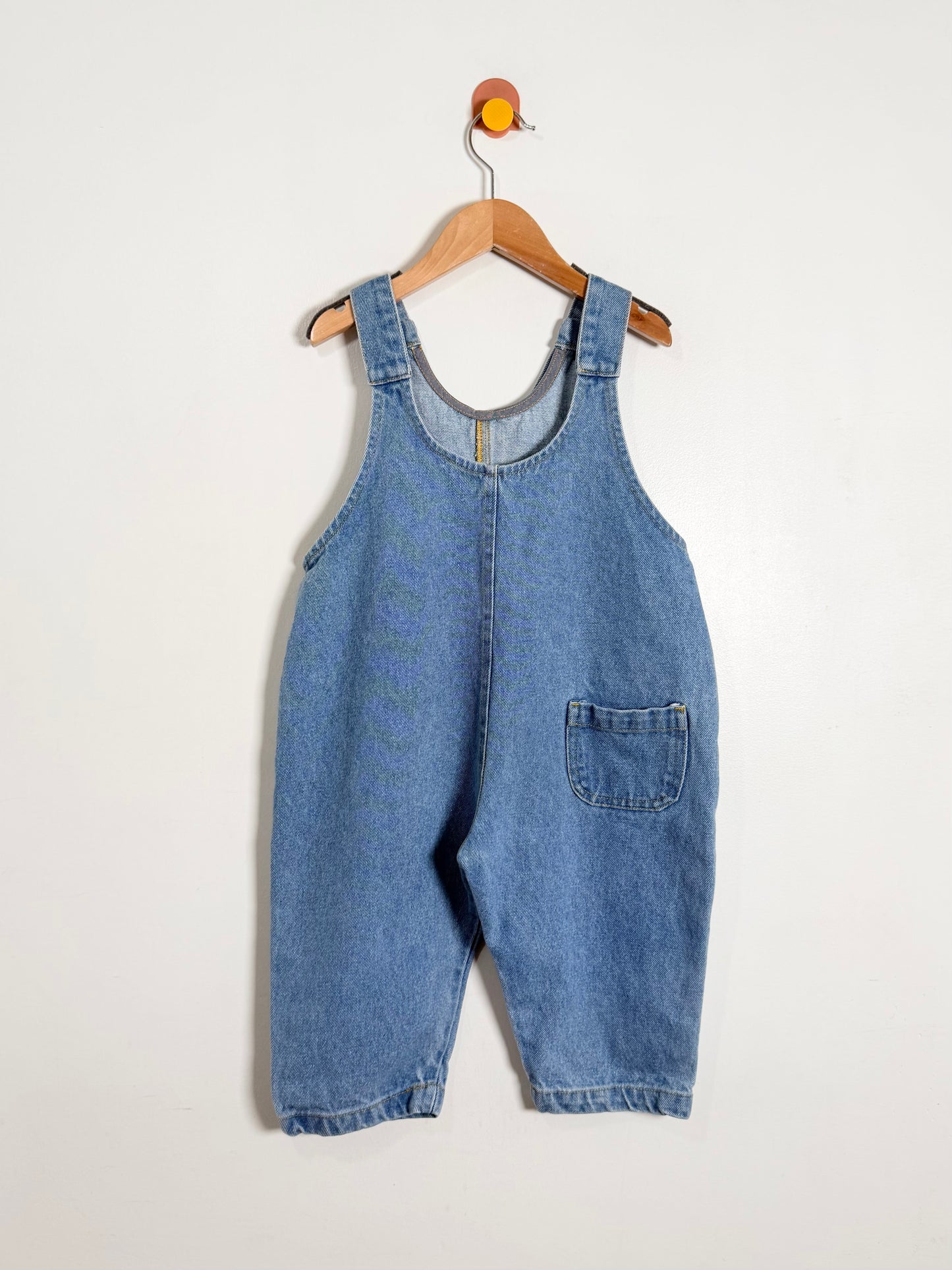 Blue denim overalls on a wooden hanger against a white background