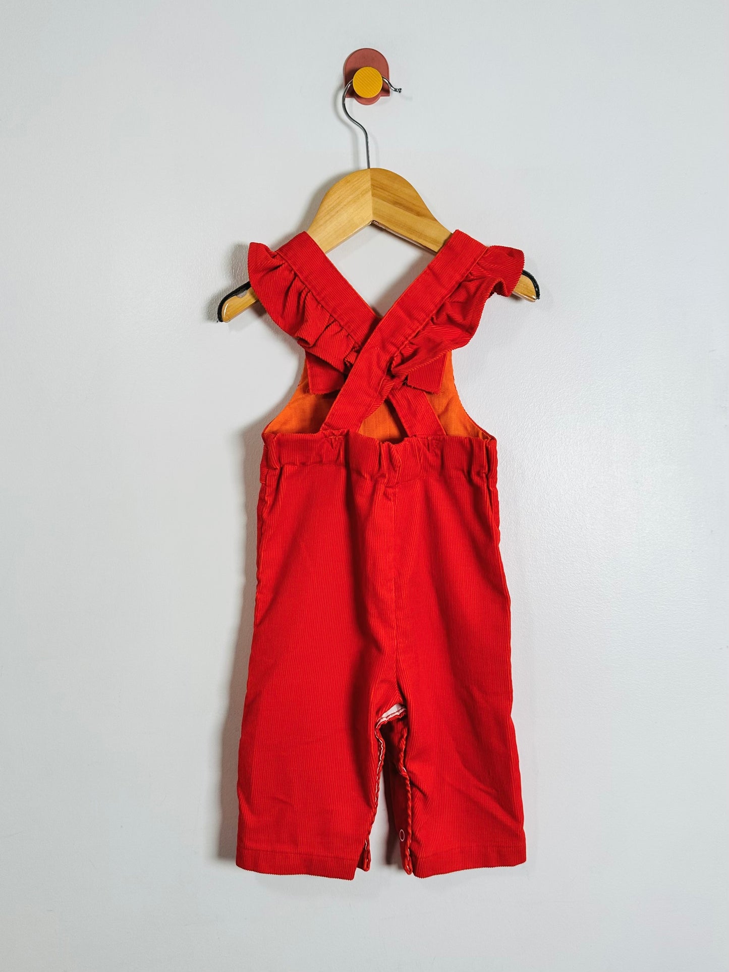 A baby romper in bright red fabric, featuring ruffled shoulder straps and a bib-style front, hanging on a wooden hanger against a plain light gray wall.