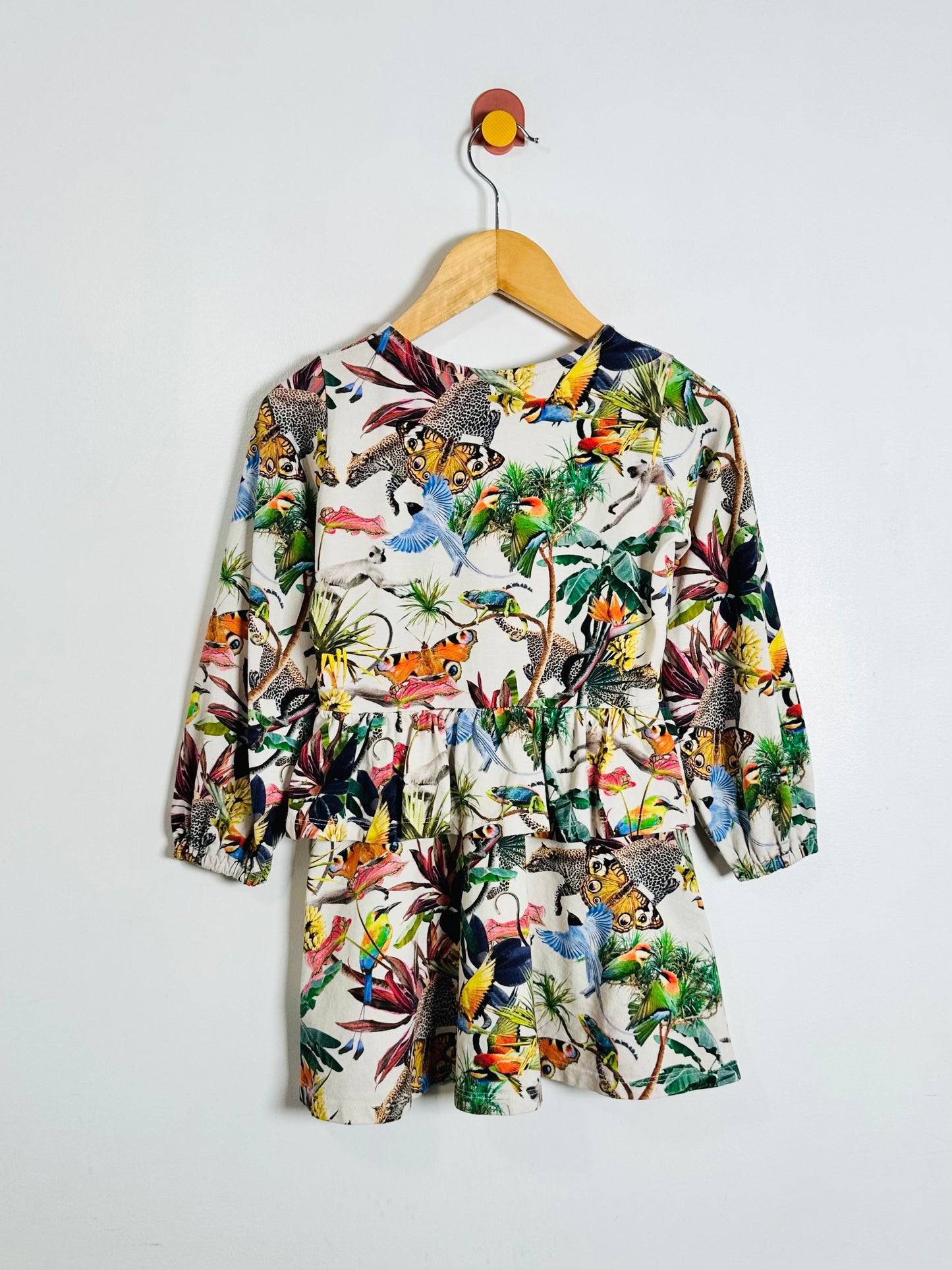 A long-sleeve children's dress with a vibrant tropical jungle print featuring toucans, monkeys, and palm trees, hanging on a wooden hanger against a plain white wall.