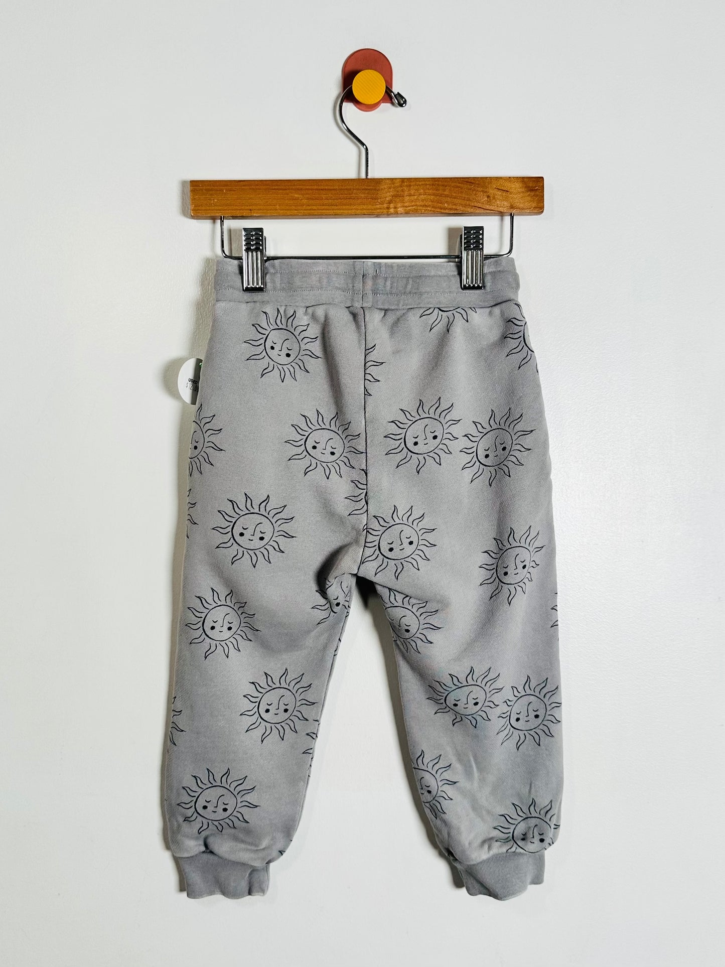 Gray jogger pants with a repeating black sun face pattern, hanging from a wooden hanger with metal clips against a plain white wall.
