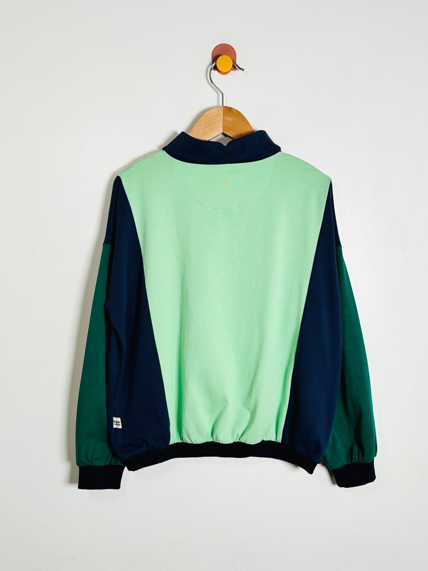 A colorblock pullover jacket hanging on a wooden hanger against a plain white wall. The back of the jacket features a light mint green center panel with dark navy blue side panels and sleeves, and a dark green panel on the right sleeve.