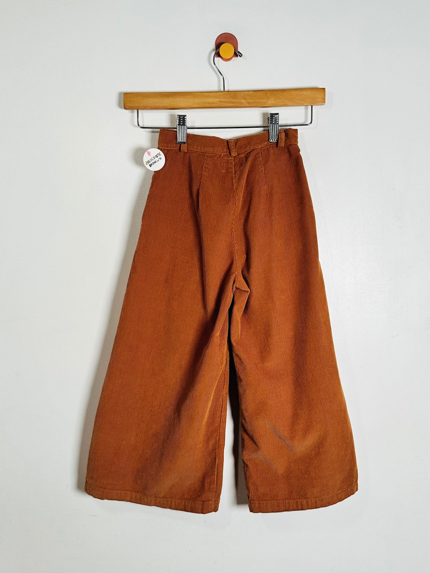 A pair of wide-leg pants in rust orange corduroy fabric, hanging from a wooden hanger against a plain off-white wall.
