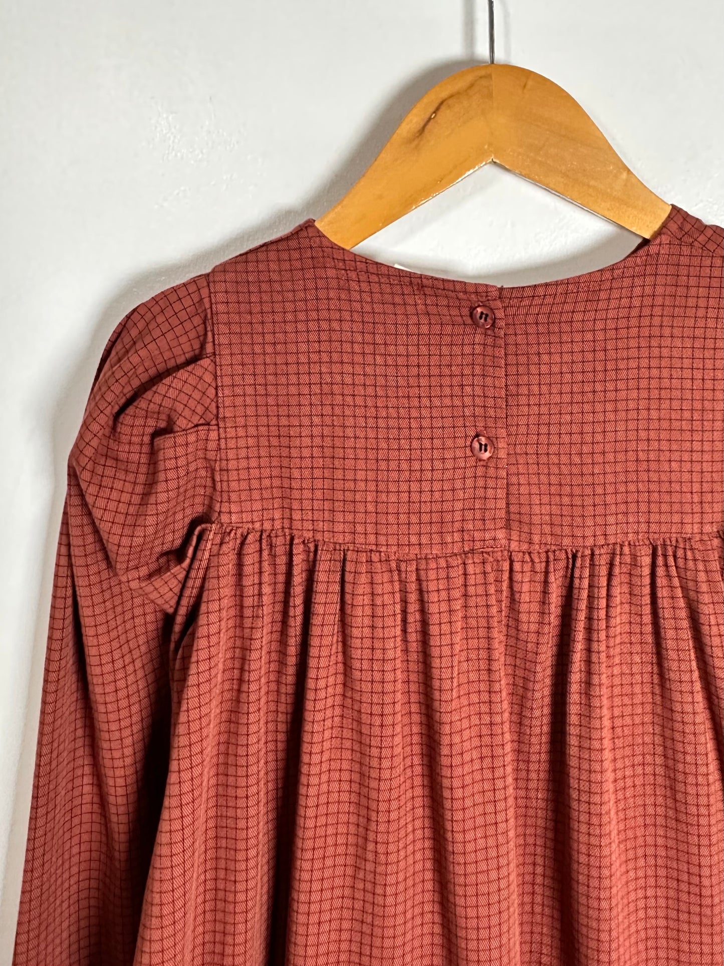 A rust-colored long-sleeve top with a small black grid pattern, hanging on a wooden hanger against a white wall.
