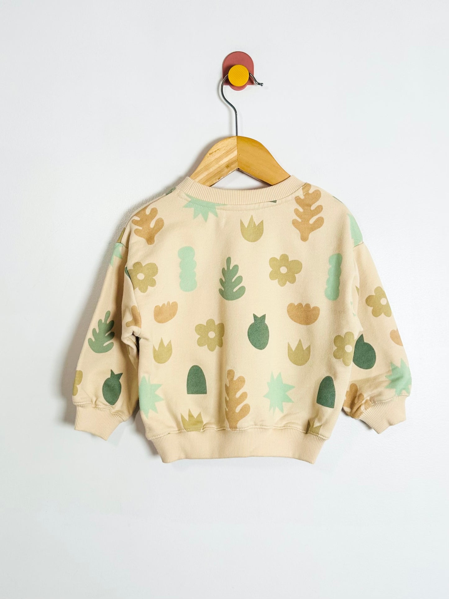 Beige sweatshirt with green leaf patterns on a hanger against a white background