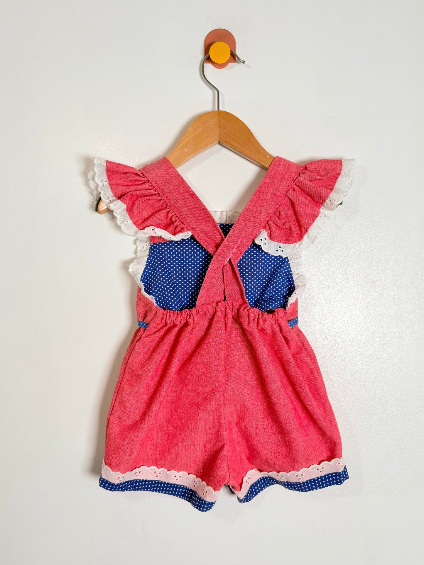 A baby girl's shortall romper in pink and blue gingham fabric, featuring ruffled cap sleeves with white lace trim, a blue polka dot bib panel, and a crossover back. The romper is displayed on a wooden hanger against a plain white wall.