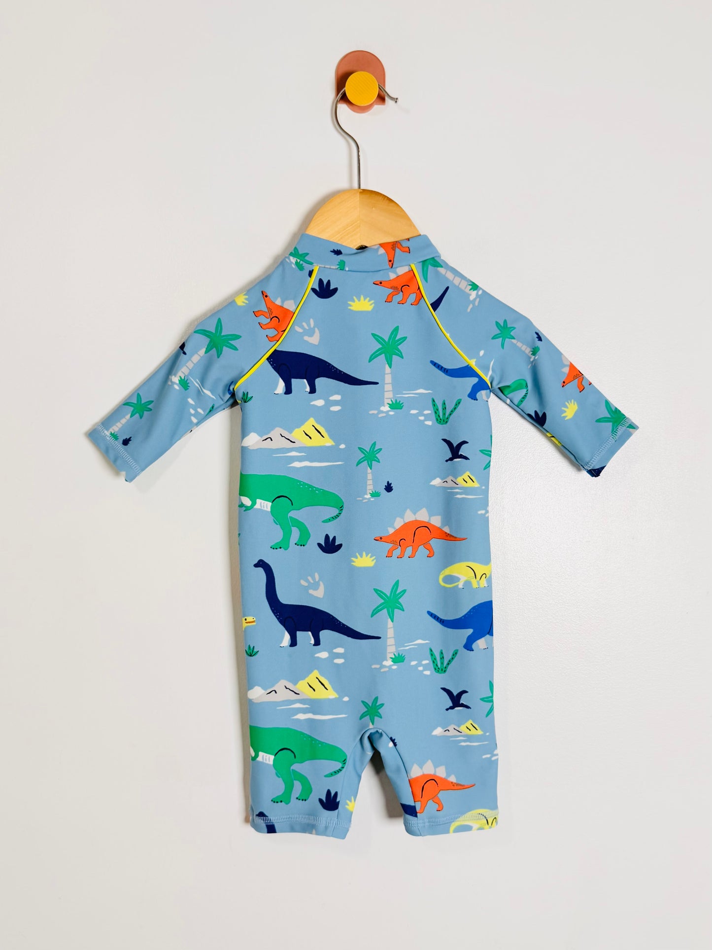 A baby long-sleeve swimsuit or one-piece in light blue, hanging on a wooden hanger against a plain light gray wall. The fabric is covered in a colorful dinosaur and beach-themed print including green, blue, orange, and yellow dinosaurs, palm trees, waves, and suns.