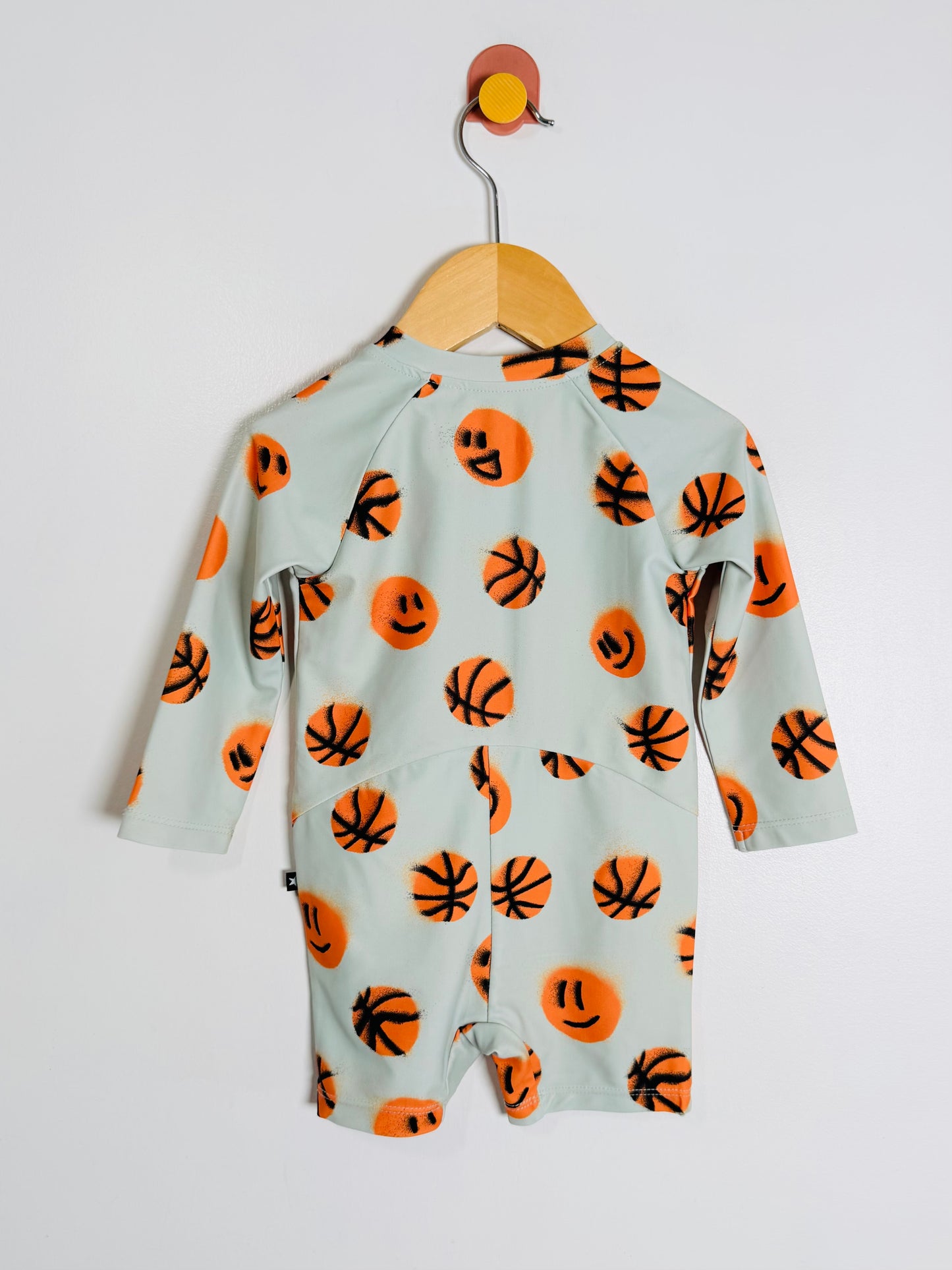 A baby long-sleeve romper in a pale sage green, featuring an all-over print of orange basketballs and smiley faces, hanging on a wooden hanger against a plain light gray wall.