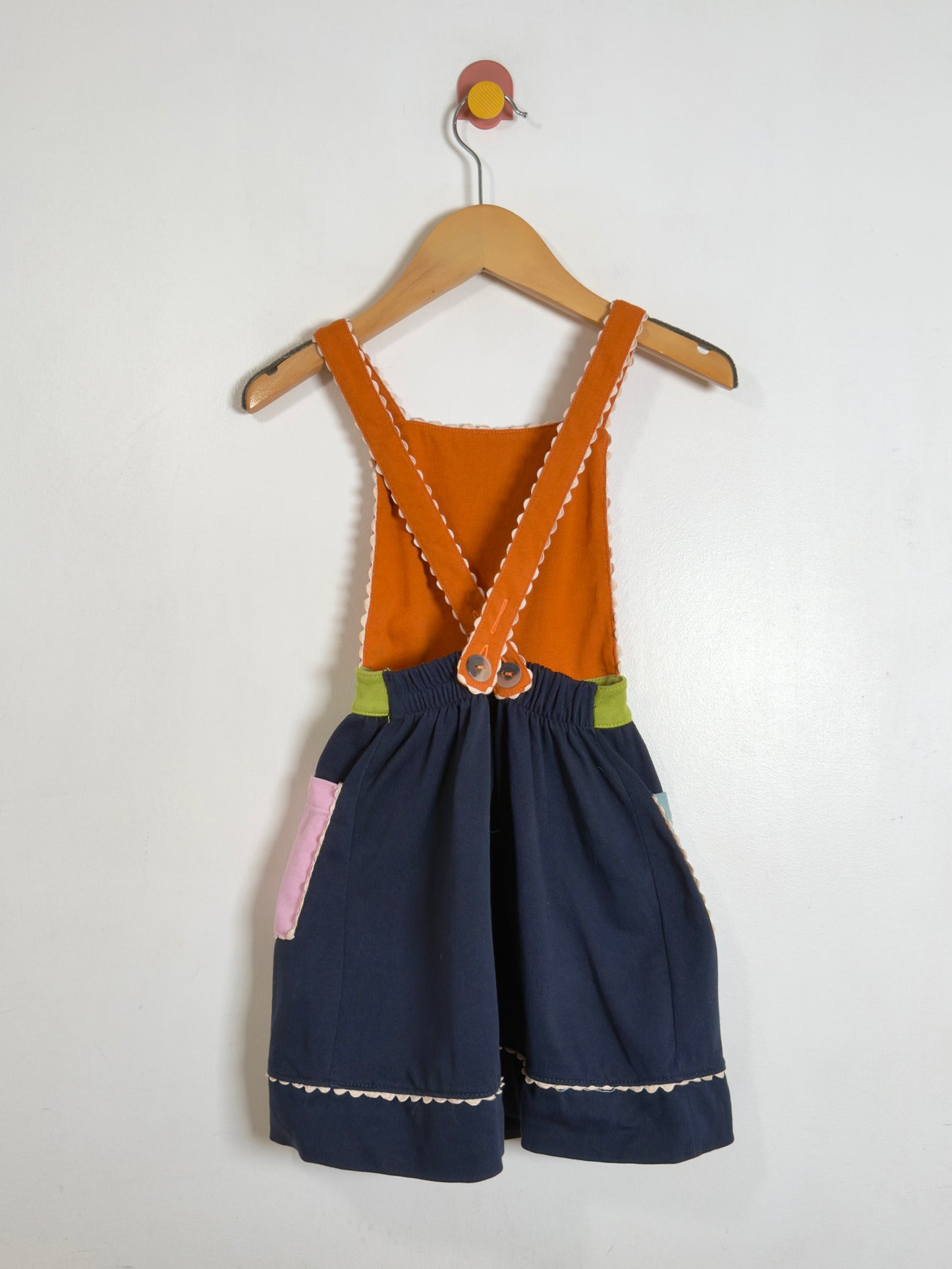 A children's sleeveless romper or short overall hanging on a wooden hanger against a plain white wall. The garment has an orange-brown bib section with wide shoulder straps, dark navy shorts, and small accent patches in light green and pink, with white contrast stitching.