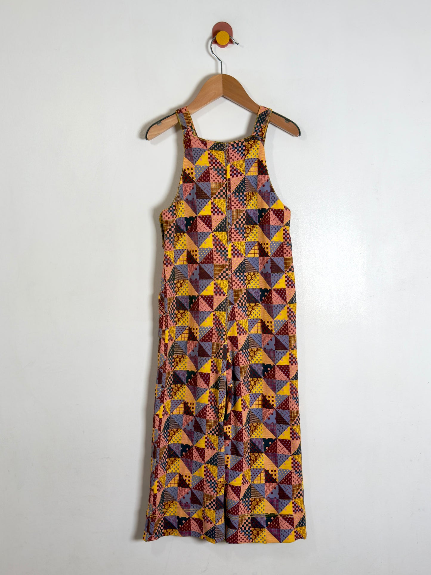 Overalls with a colorful geometric patchwork print, hanging on a wooden hanger against a plain white wall.