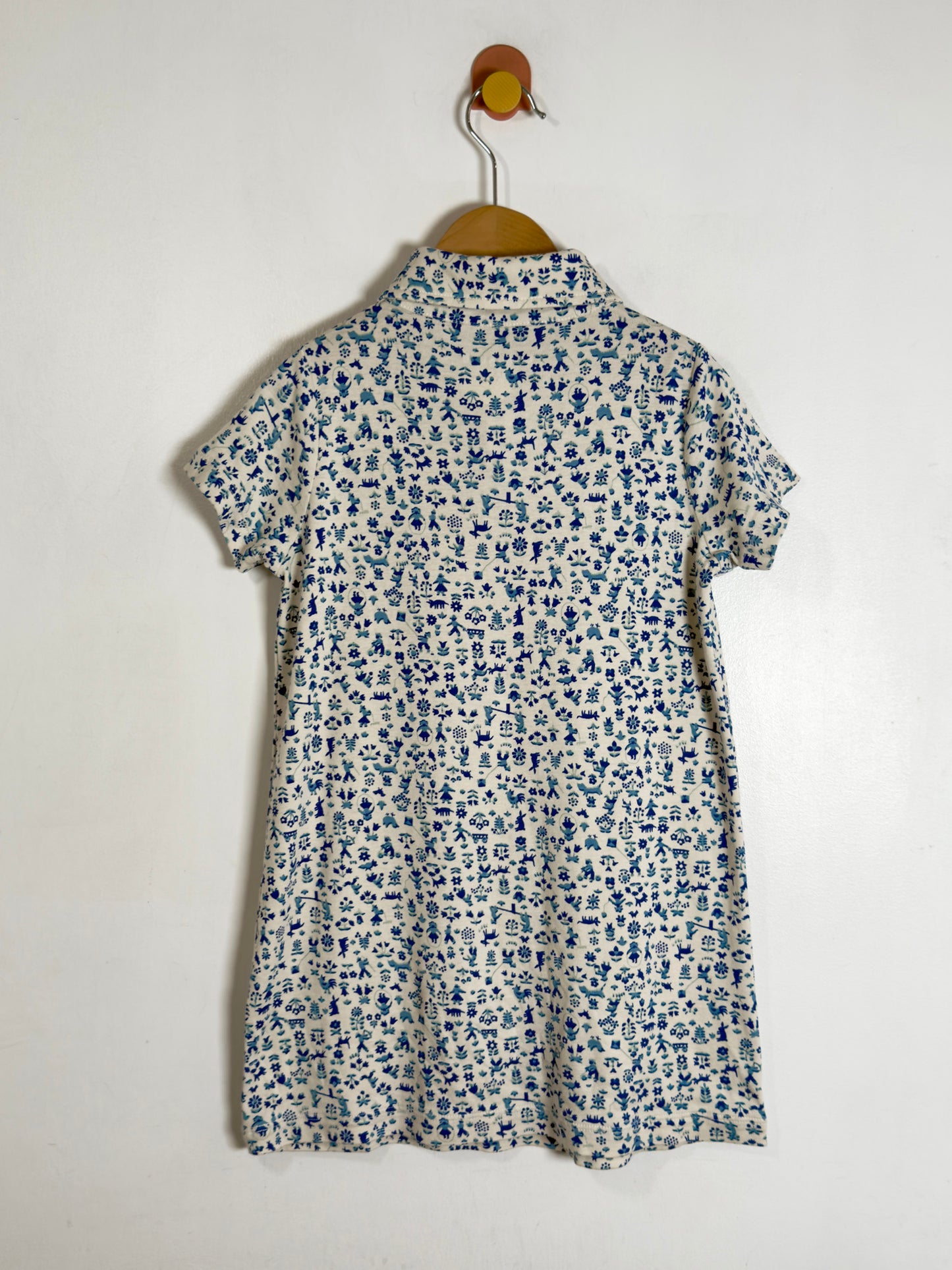 A short-sleeve collared shirt dress with a dense blue and white floral print, hanging on a wooden hanger against a plain off-white wall.
