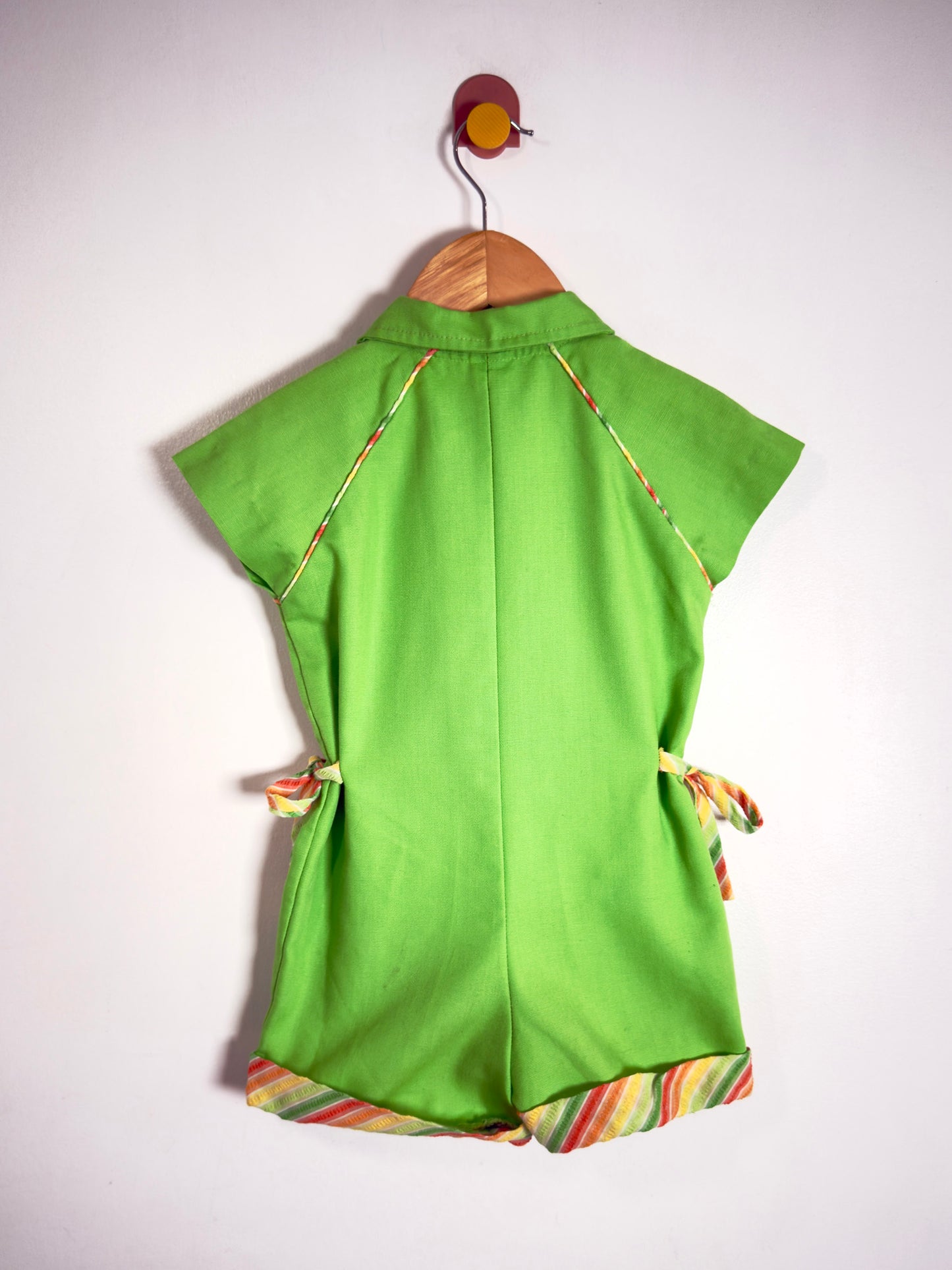 A bright green short-sleeve romper or jumpsuit hanging on a wooden hanger against a plain white wall. The garment has thin multicolor striped piping details along the shoulders and a matching multicolor striped trim along the hem, with side ties.
