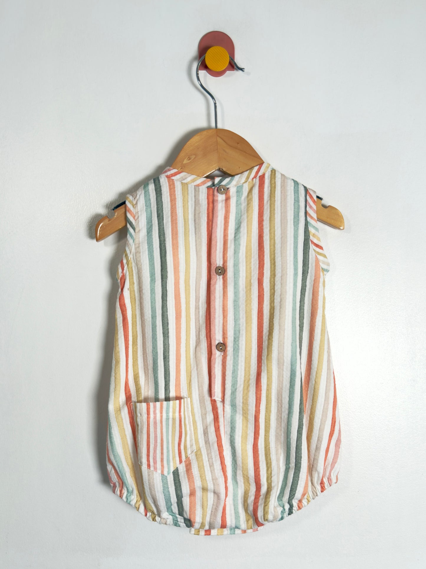 A sleeveless button-front baby top with vertical stripes in orange, green, yellow, and white, hanging on a wooden hanger against a plain white wall.