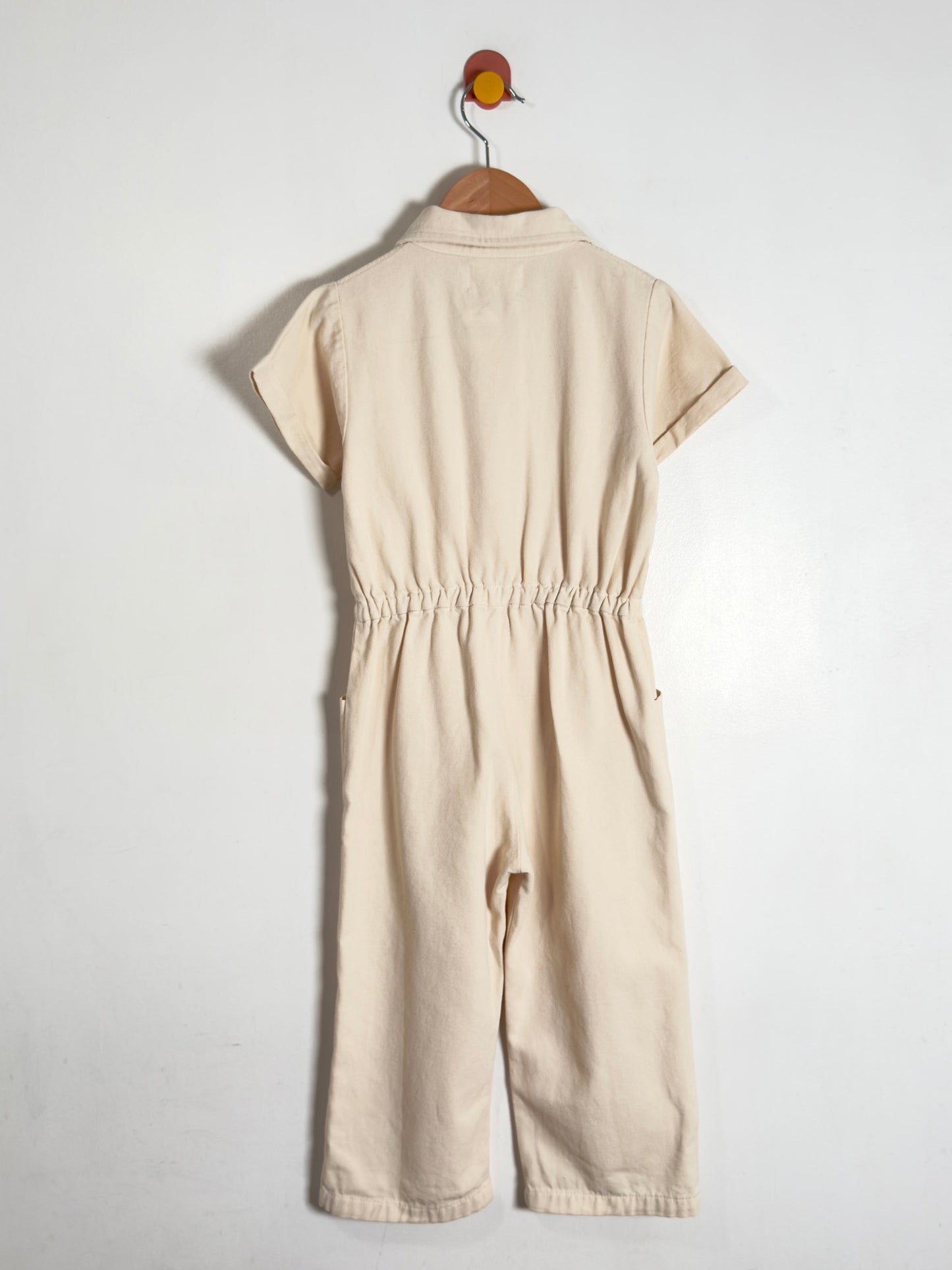 A light beige short-sleeve jumpsuit with an elastic waist, hanging on a wooden hanger against a plain white wall.