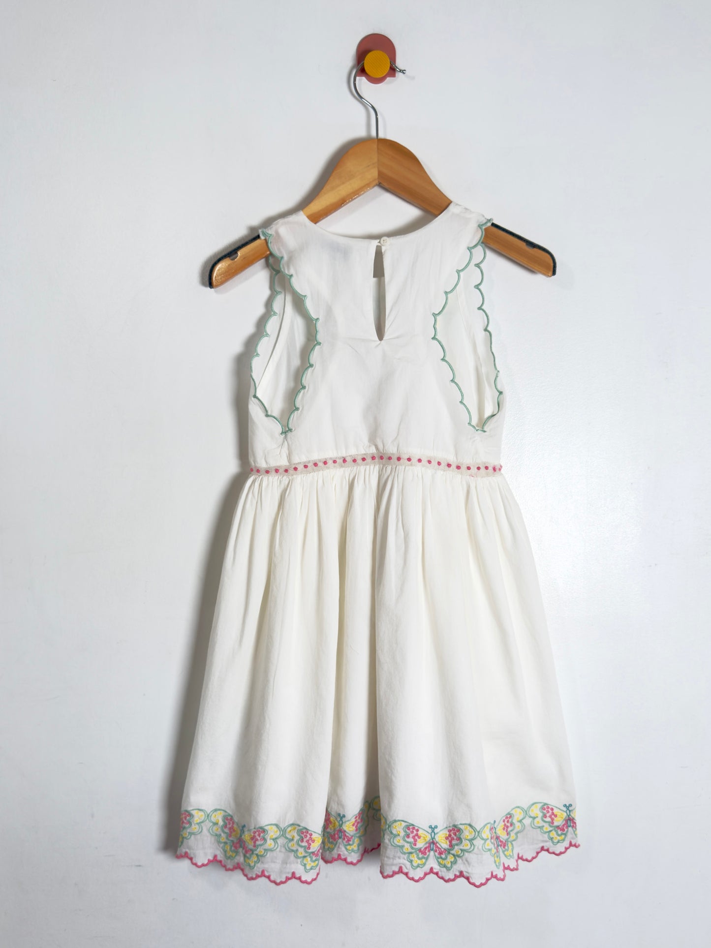An off-white sleeveless baby dress with green embroidered details and a floral lace hem, hanging on a wooden hanger against a plain white wall.