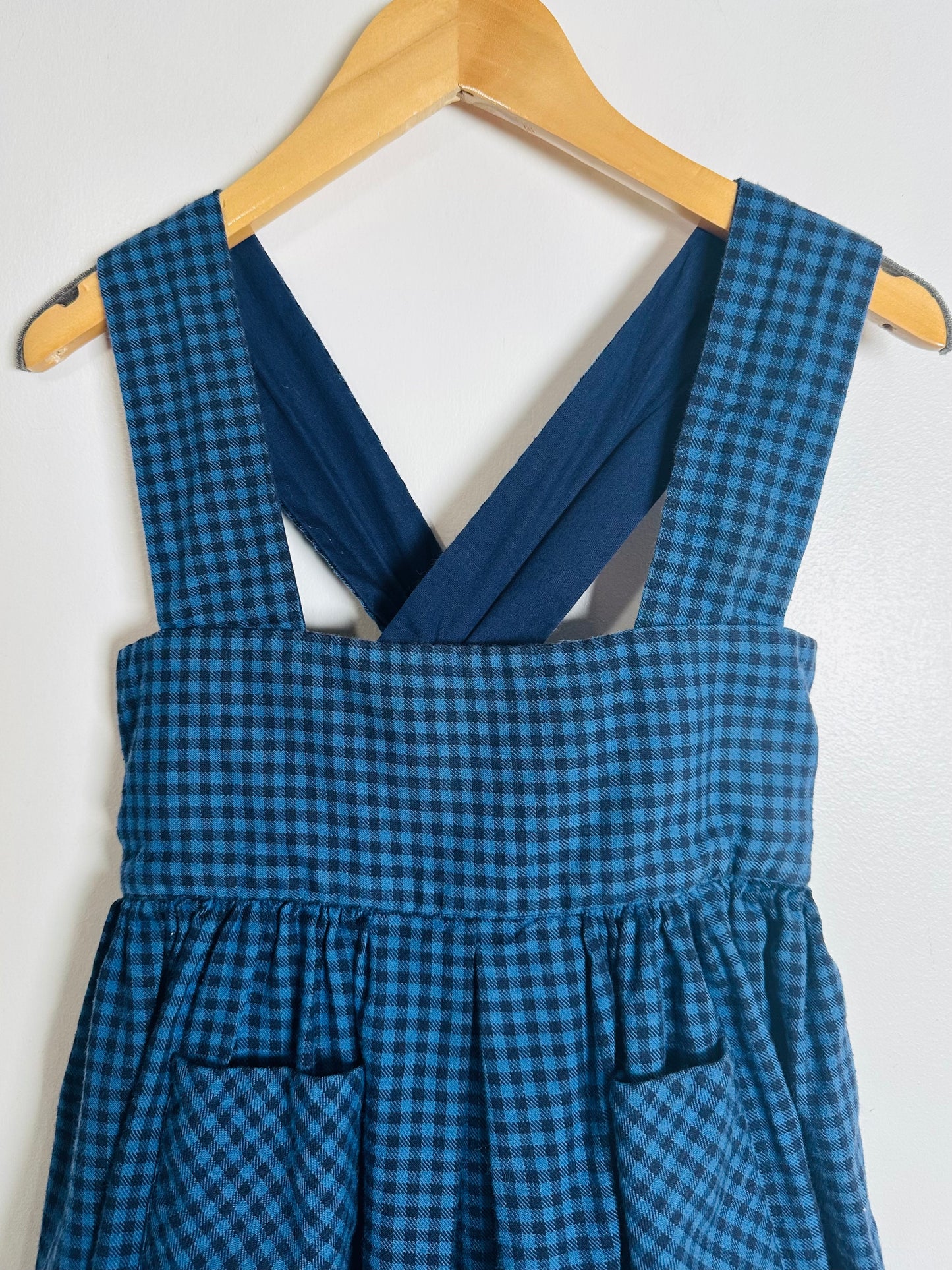 Blue checkered pinafore dress on a wooden hanger against a white background