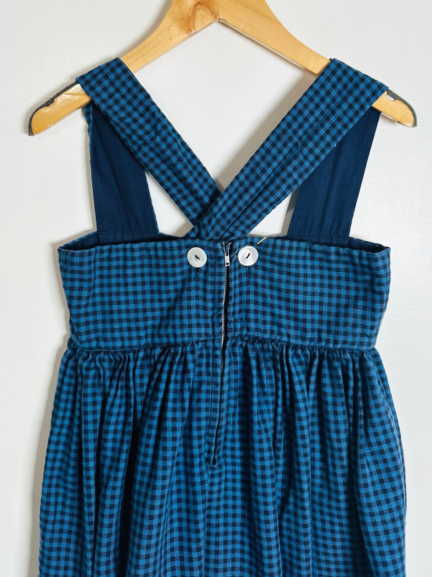 Blue checkered dress with suspenders on a wooden hanger against a white background