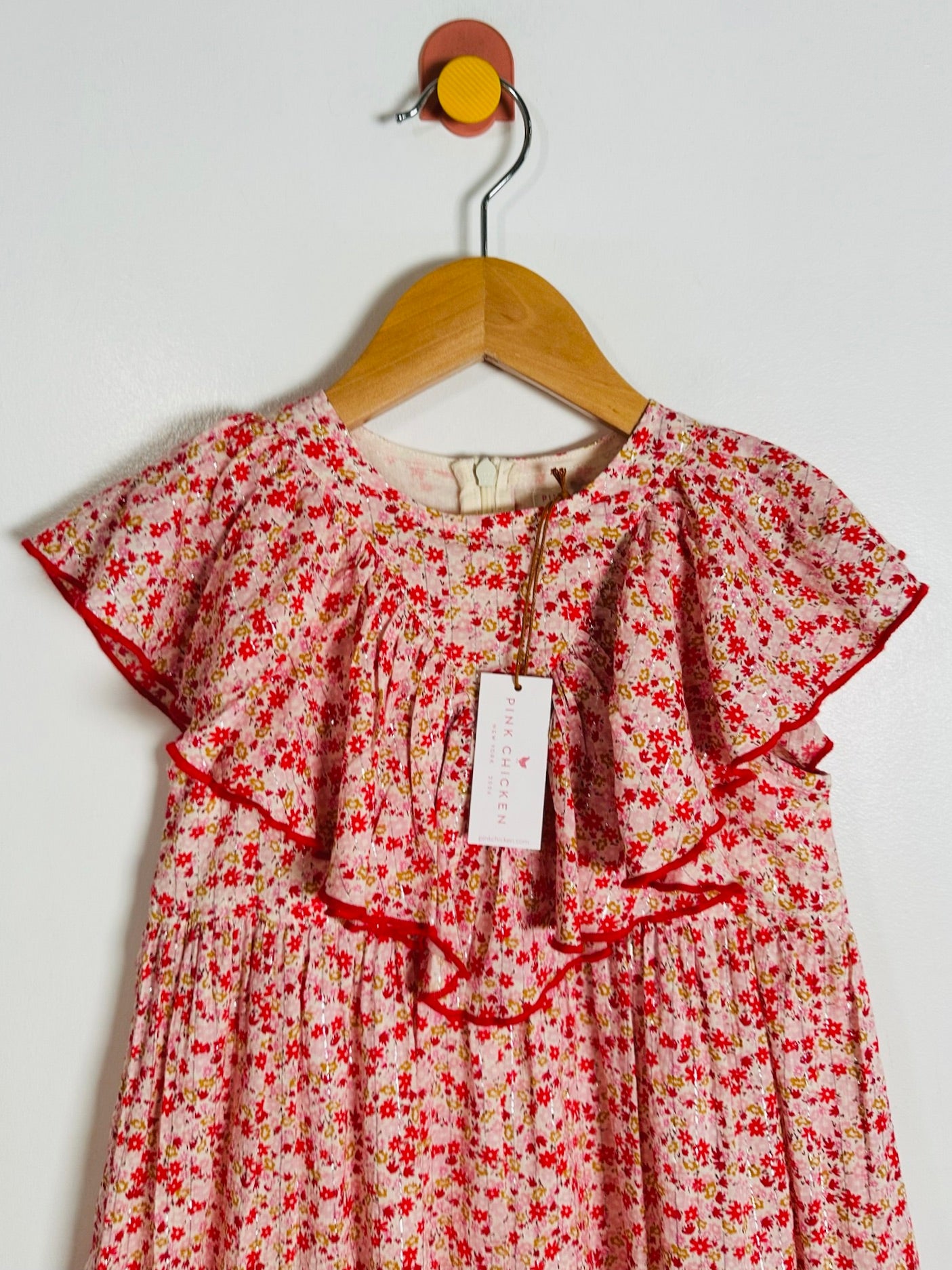 A pink short-sleeve baby dress with a red and yellow floral print, featuring ruffled trim and a hanging tag, displayed on a wooden hanger against a plain white wall.