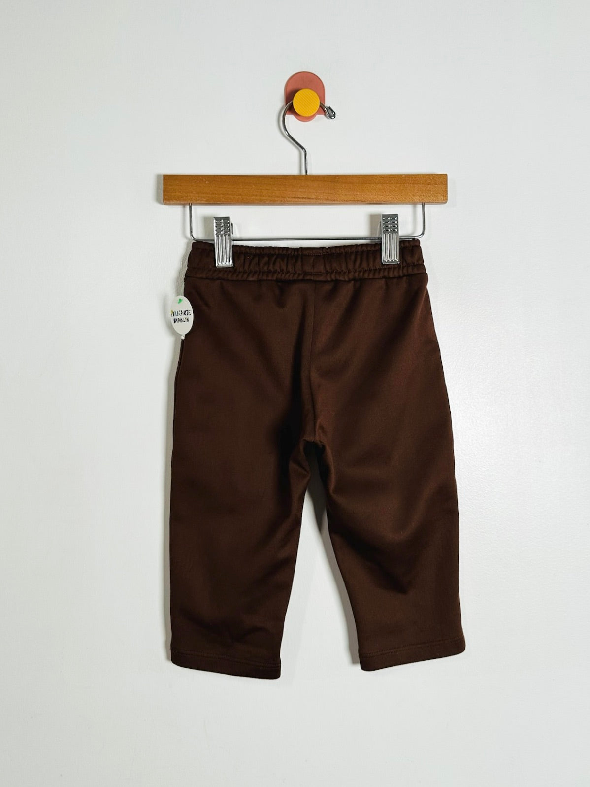 A pair of toddler brown capri pants hanging from a wooden hanger against a plain white wall.
