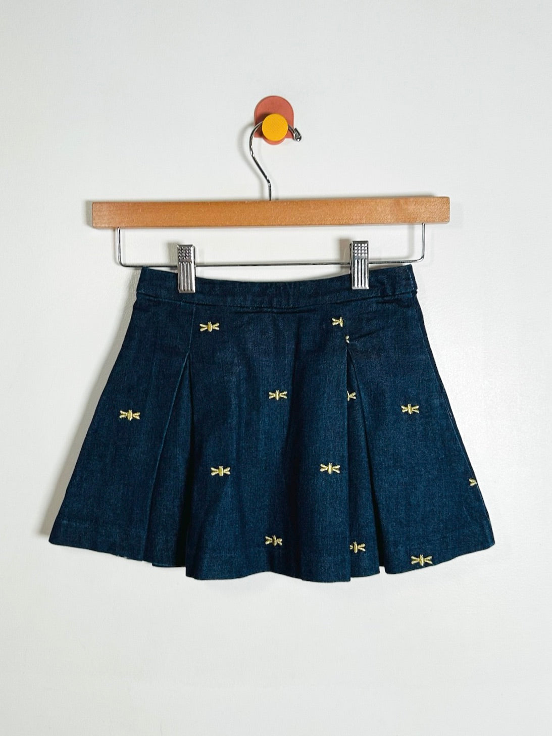 A dark blue pleated mini skirt with small yellow embroidered motifs, hanging from a wooden hanger against a plain white wall.