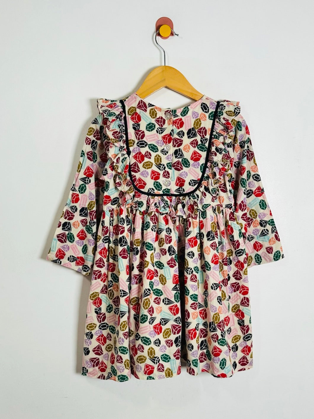 A long-sleeve dress with a colorful gem print on a light pink background, hanging on a wooden hanger against a plain white wall.