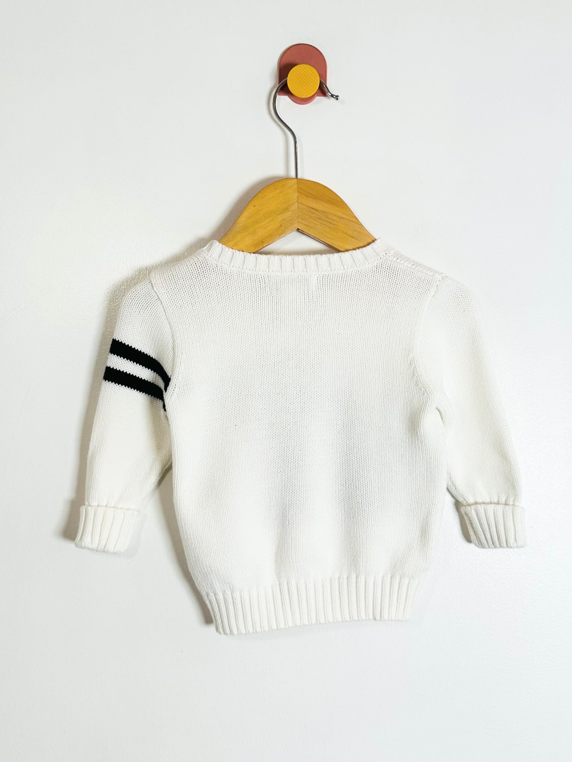 White sweater with black stripes on sleeves hanging on a wooden hanger against a white background