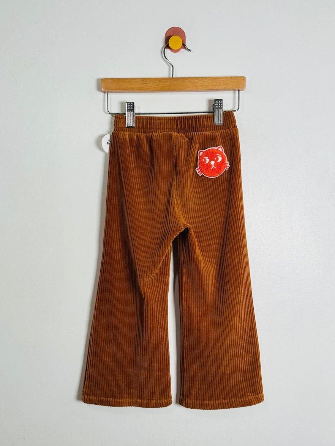 A pair of brown corduroy wide-leg pants hanging from a wooden hanger against a plain white wall. A red cartoon sticker is applied to the left thigh.