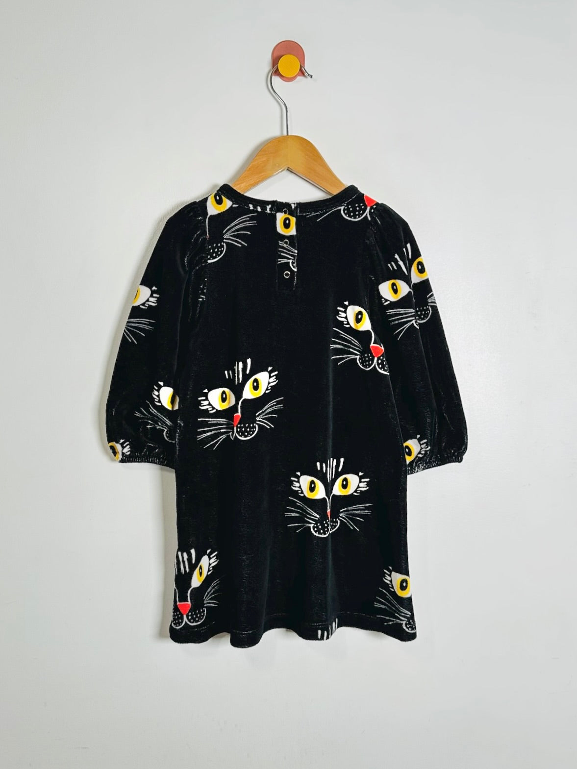 A black velvet long-sleeve dress with a playful cat face print, hanging on a wooden hanger against a plain white wall.