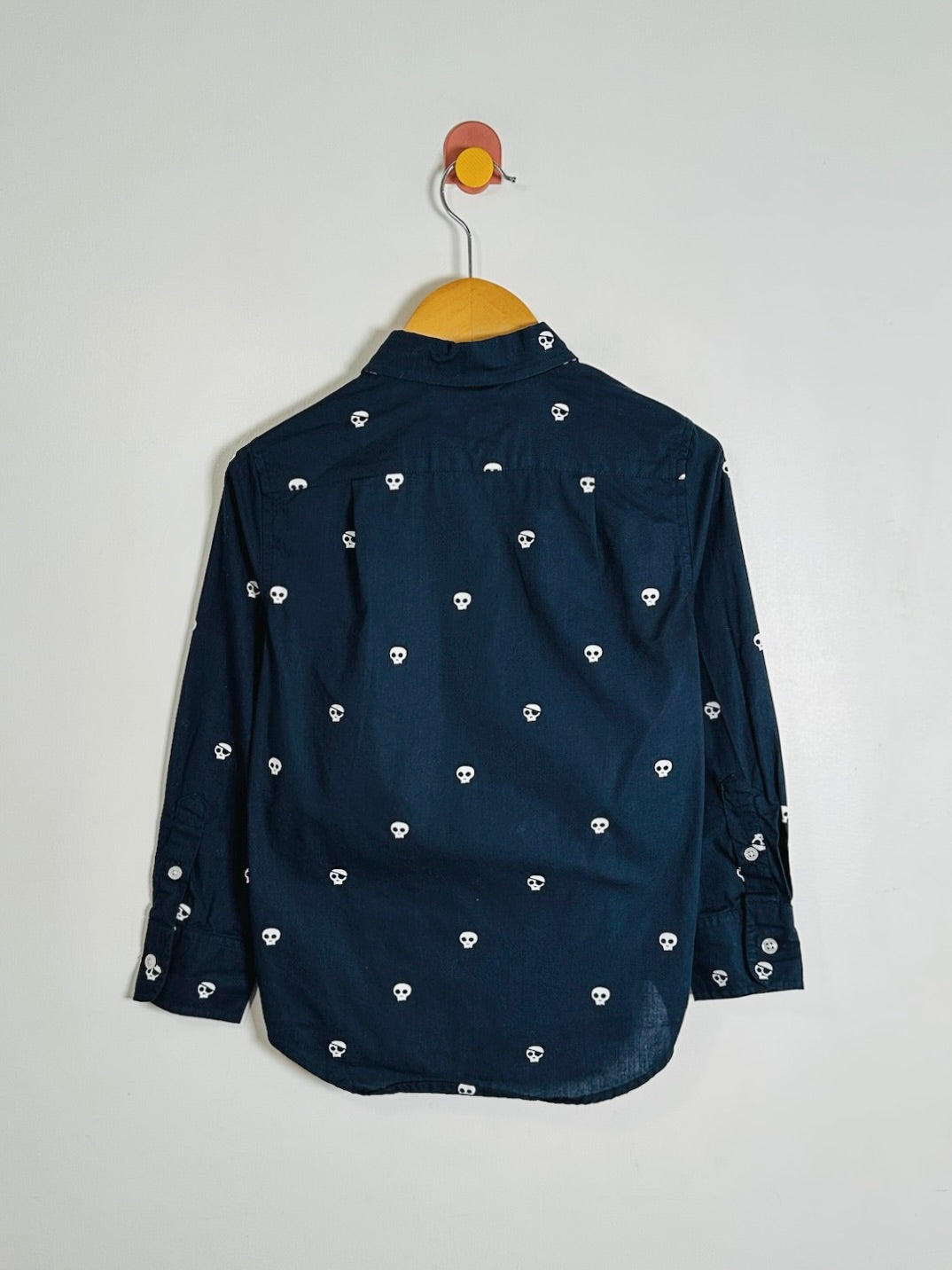 A dark navy blue long-sleeve button-up shirt with a repeating white skull pattern, hanging on a wooden hanger against a plain light gray wall.