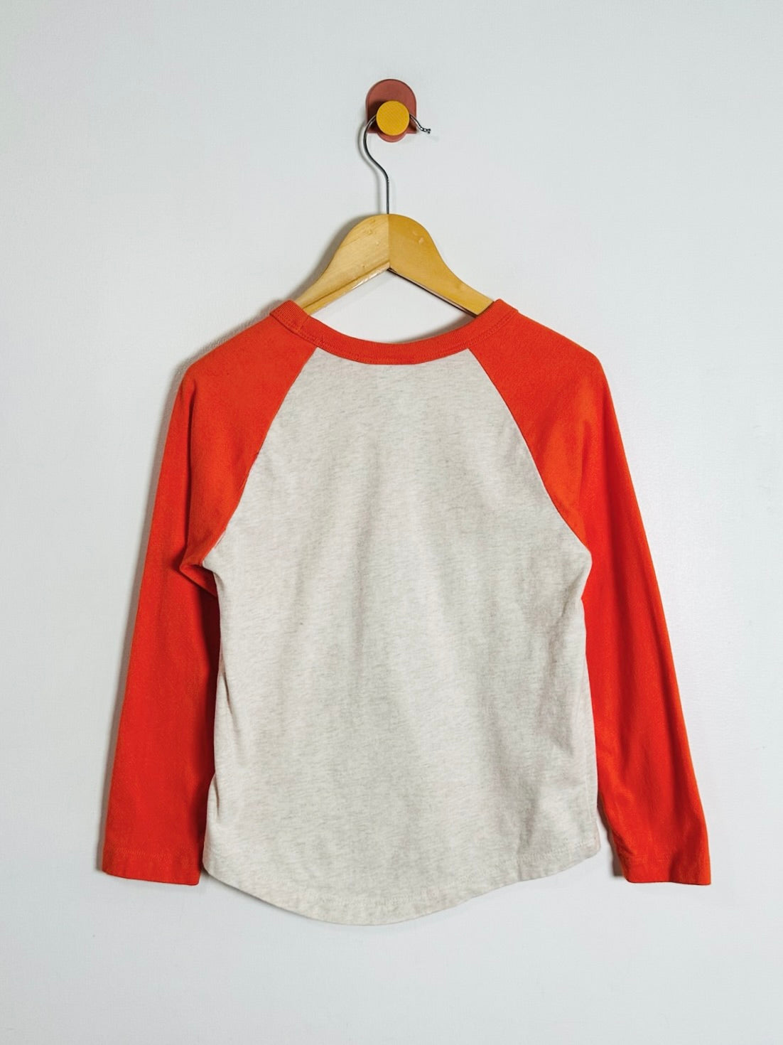 A raglan baseball-style long-sleeve shirt hanging on a wooden hanger against a plain light gray wall. The shirt has a light heather gray body and bright red sleeves and collar.