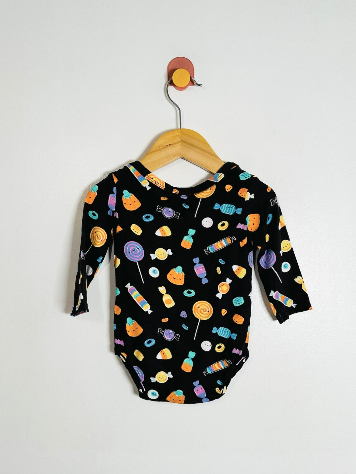 A baby long-sleeve bodysuit with a colorful candy print on a black background, hanging on a wooden hanger against a plain white background.