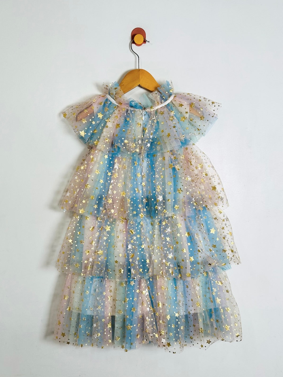 A tiered tulle dress for toddlers or girls, decorated with gold star-shaped sequins and iridescent pastel shimmer. The dress features a high ruffled collar and short flutter sleeves, hanging on a wooden hanger against a plain white wall.
