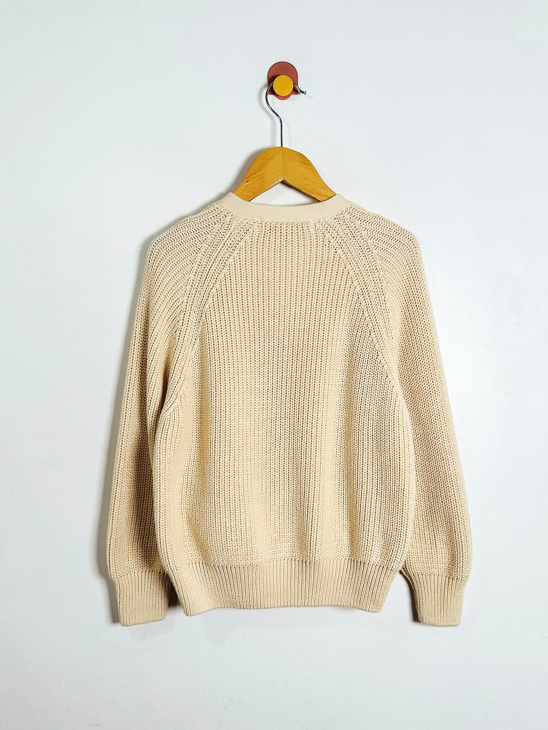 A cream-colored knit sweater with raglan sleeves hanging on a wooden hanger against a plain white wall.