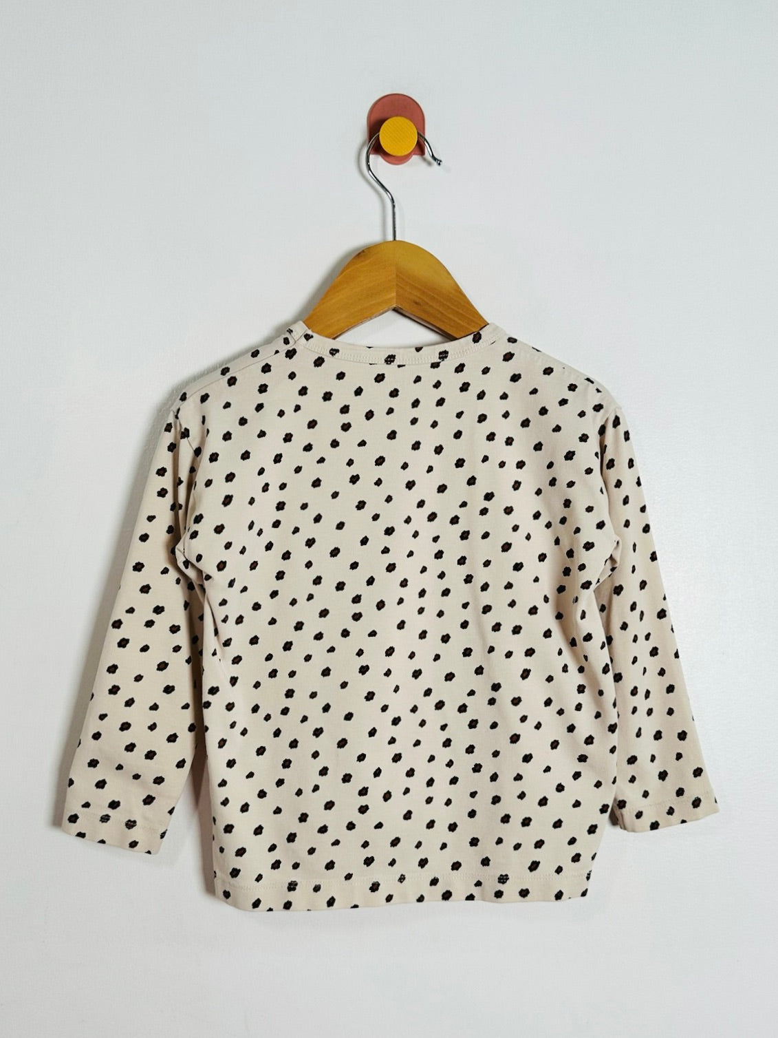 A cream-colored long-sleeve top with a black polka dot pattern, hanging on a wooden hanger against a plain white wall.