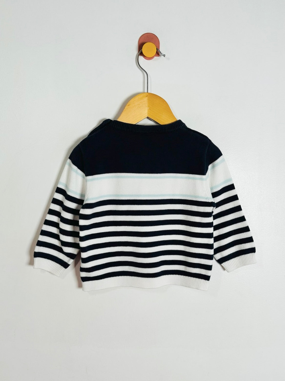 A baby knit sweater with a navy blue yoke and navy and white horizontal stripes on the sleeves and body, hanging on a wooden hanger against a plain white wall.