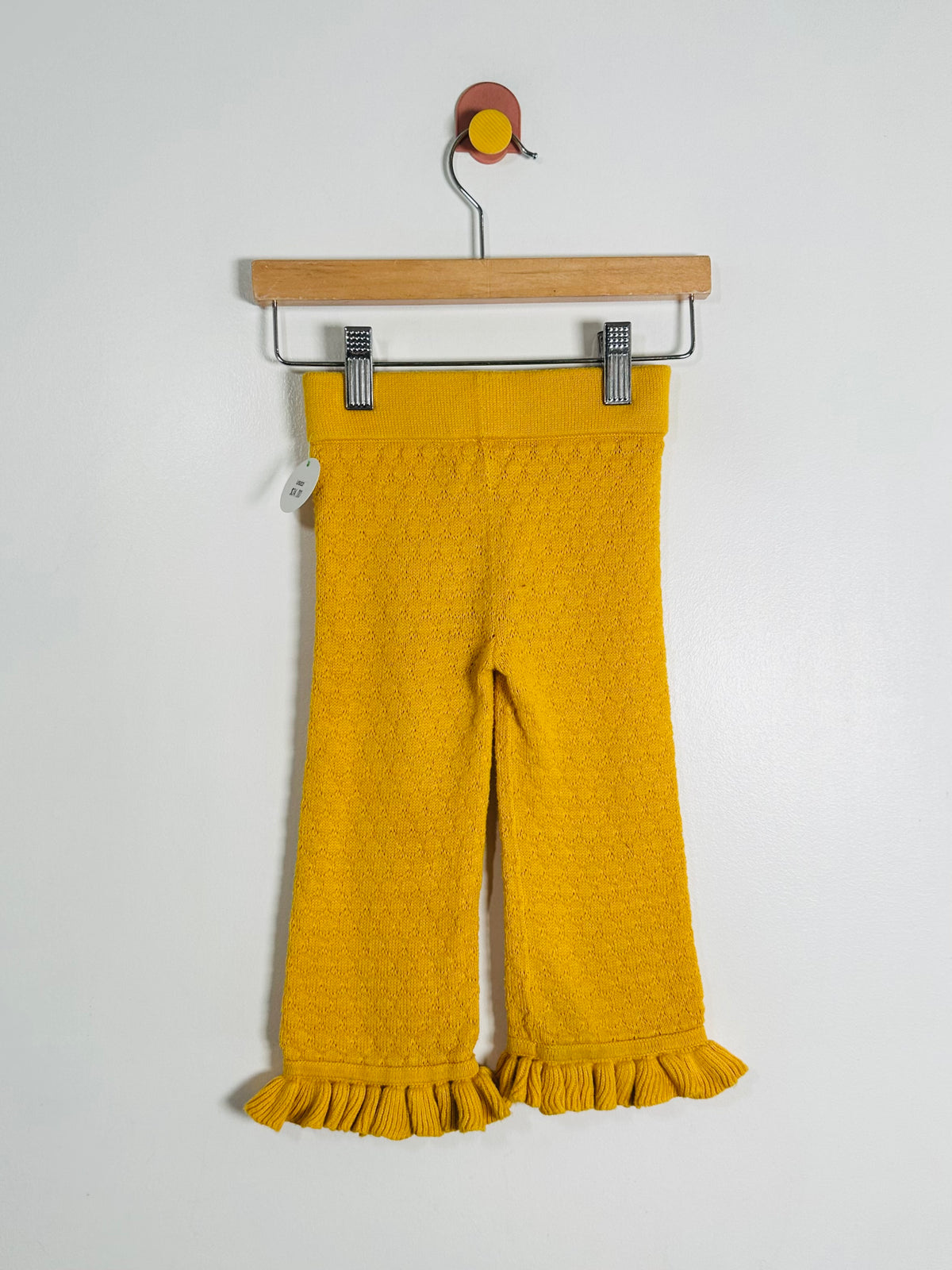 A pair of mustard yellow knit cropped pants with a textured pattern and ruffled hems, hanging from a wooden hanger against a plain white wall.
