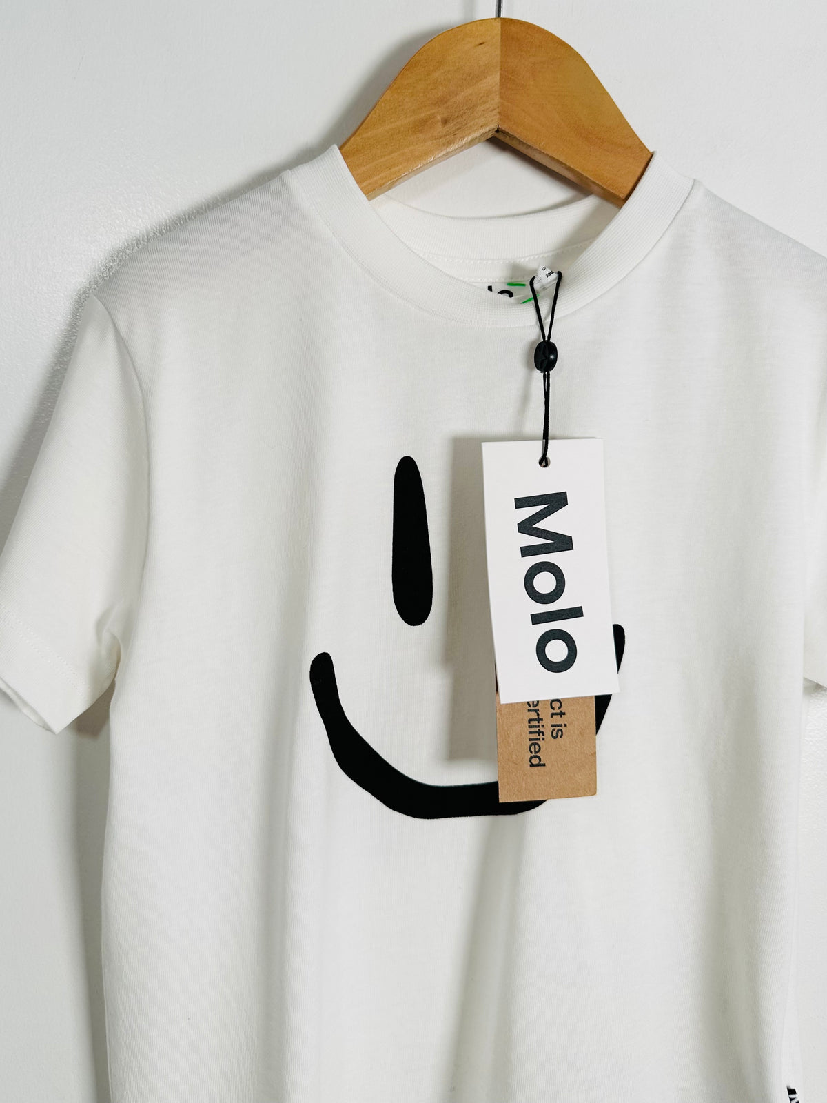 A white short-sleeve crewneck t-shirt hanging on a wooden hanger against a white wall. The shirt features a minimalist black graphic of a smiling face on the front, with a paper brand tag that reads 'Molo' attached near the collar.