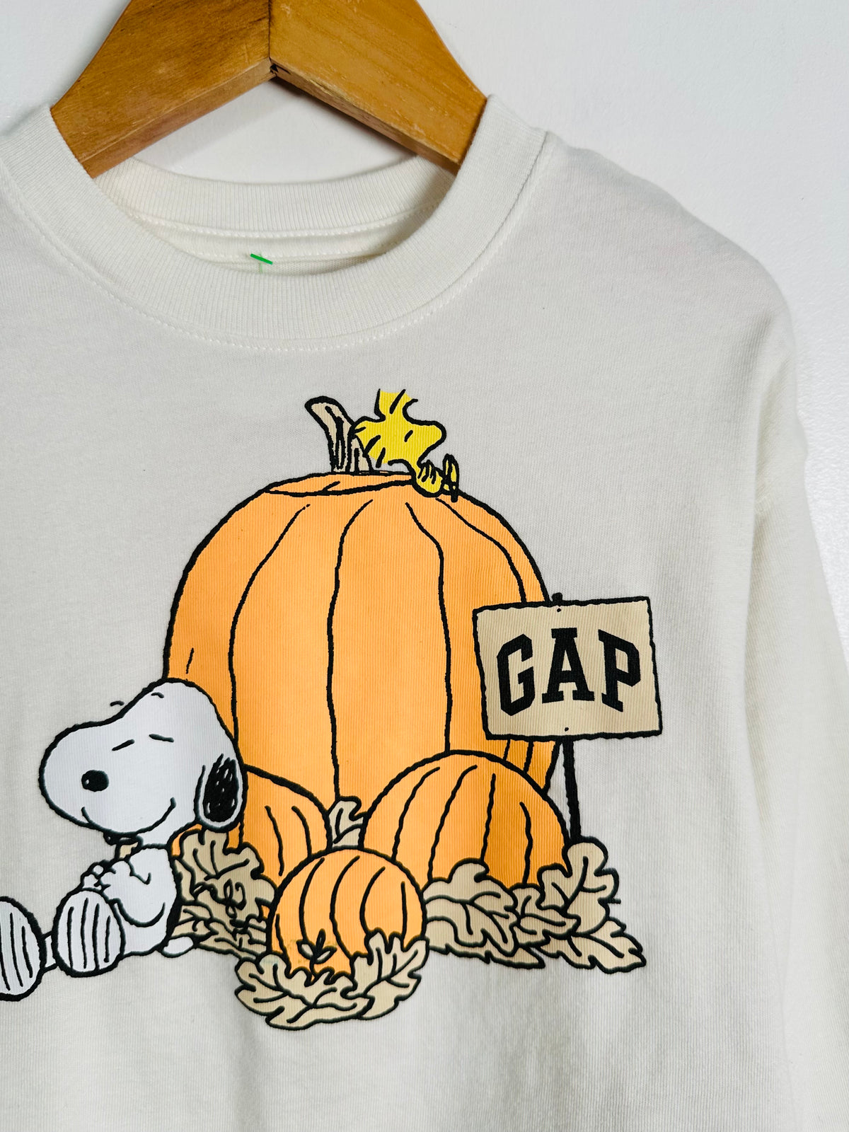 A white long-sleeve st-shirt hanging on a wooden hanger, featuring a graphic of Snoopy and Woodstock with pumpkins and a 'GAP' sign.