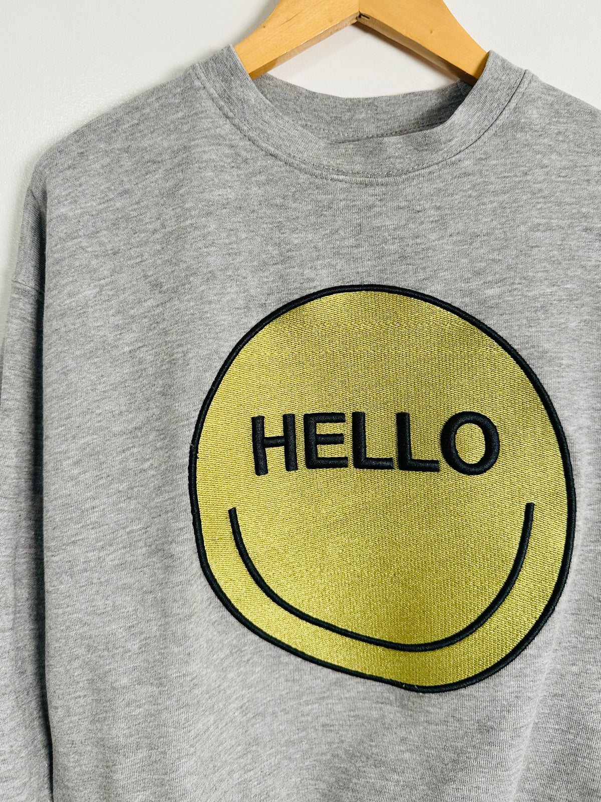 A heather gray crewneck sweatshirt hanging on a wooden hanger, featuring a large yellow smiley face graphic on the front with the word 'HELLO' embroidered in black.