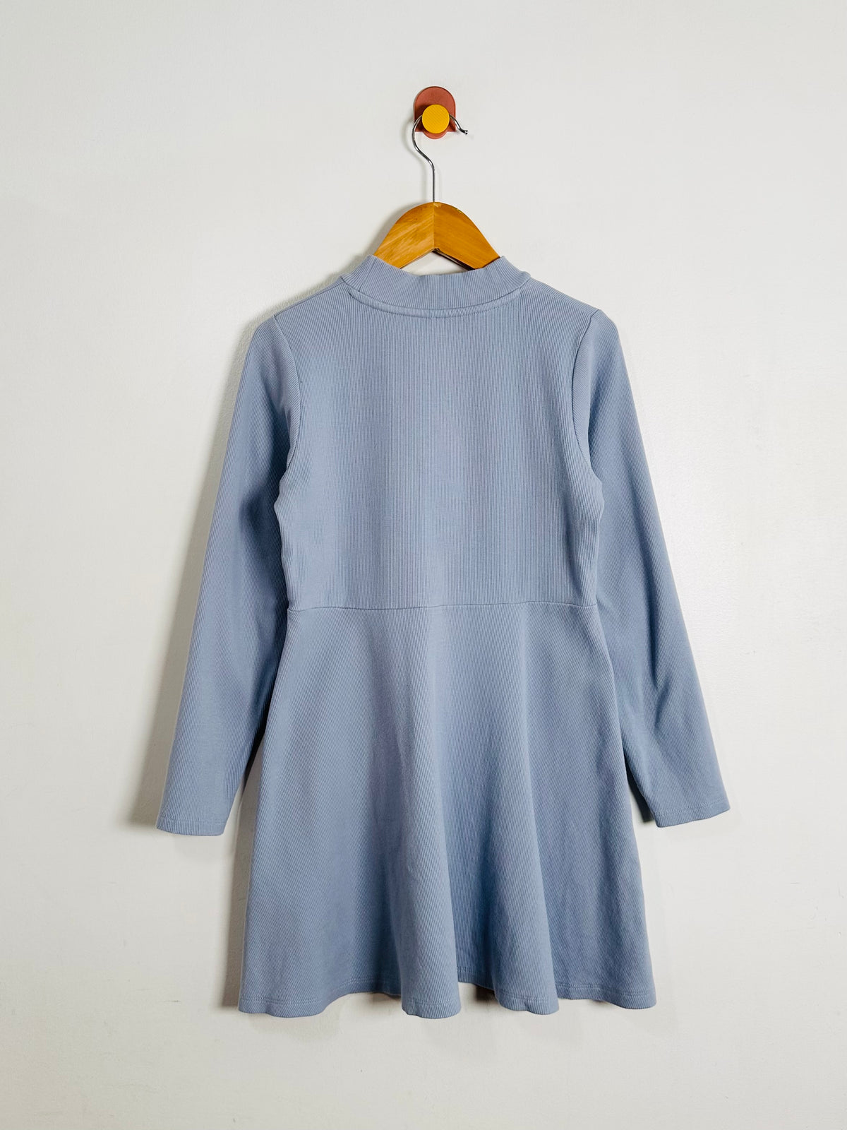 A light blue long-sleeve ribbed knit dress with a mock neck and flared skirt, hanging on a wooden hanger against a plain off-white wall.